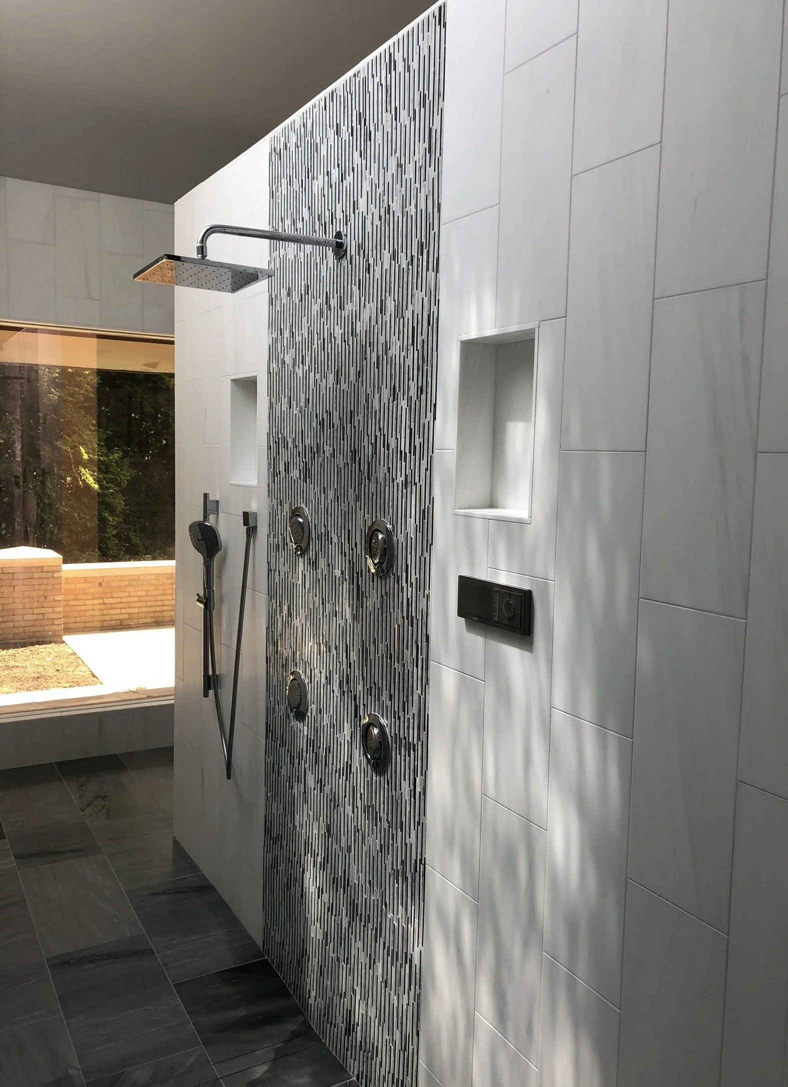 Modern outdoor shower with a rainfall showerhead, handheld shower, body jets, niche shelves, and a digital control panel, set on a tiled wall with a view of trees and a brick structure outside.