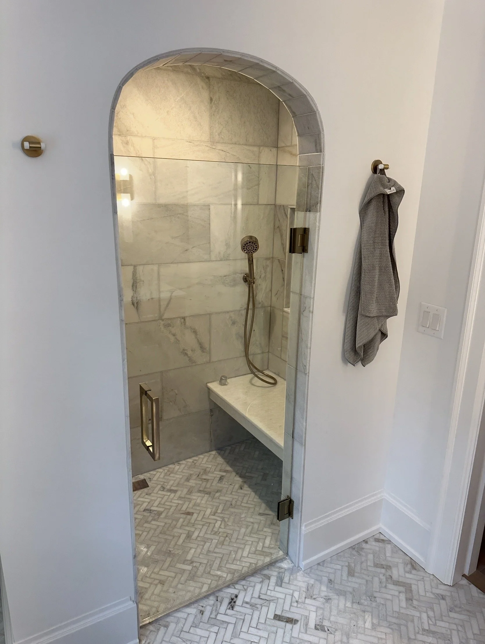 A glass-enclosed shower area with marble tile walls and a herringbone-patterned tile floor.