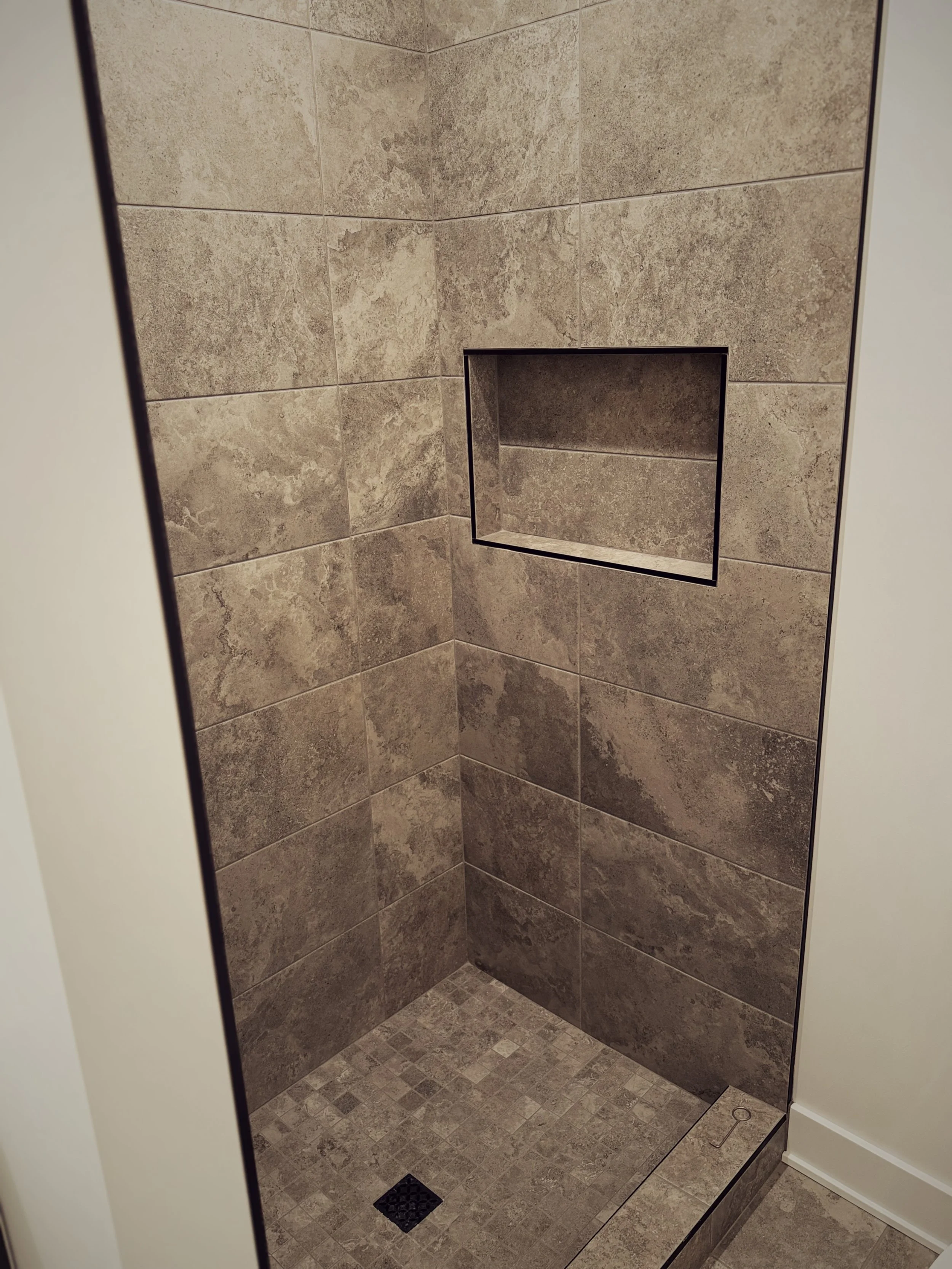 An empty walk-in shower with beige tile walls, a built-in shelf, and a black drain in the beige tiled floor.