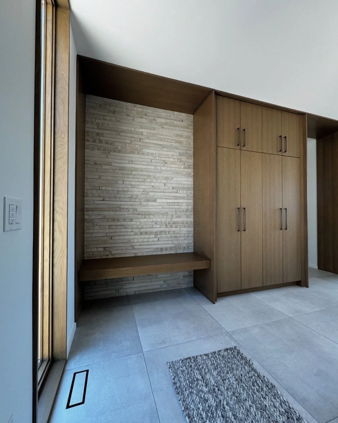 Modern home interior with wooden built-in cabinets, a bench, tile flooring, a textured wall, and a sliding glass door.