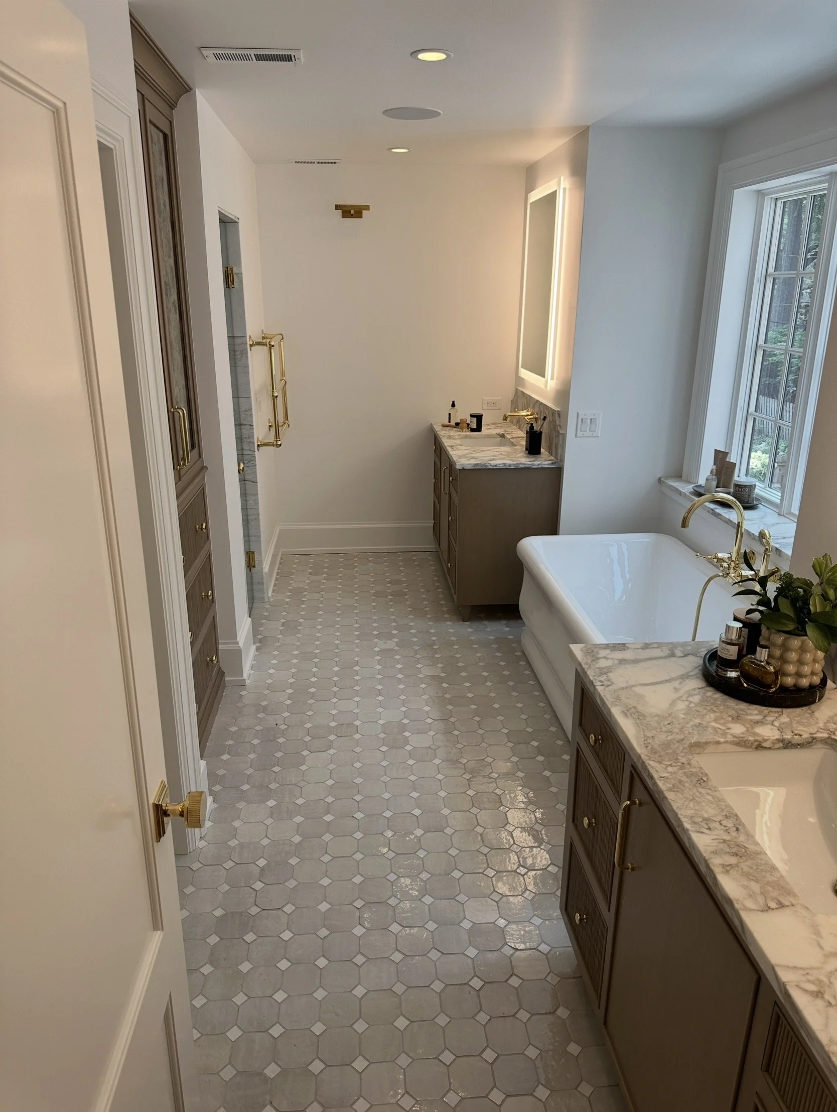 A spacious bathroom featuring a clawfoot bathtub under a large window, with marble countertops, wooden cabinetry, and a tiled floor.