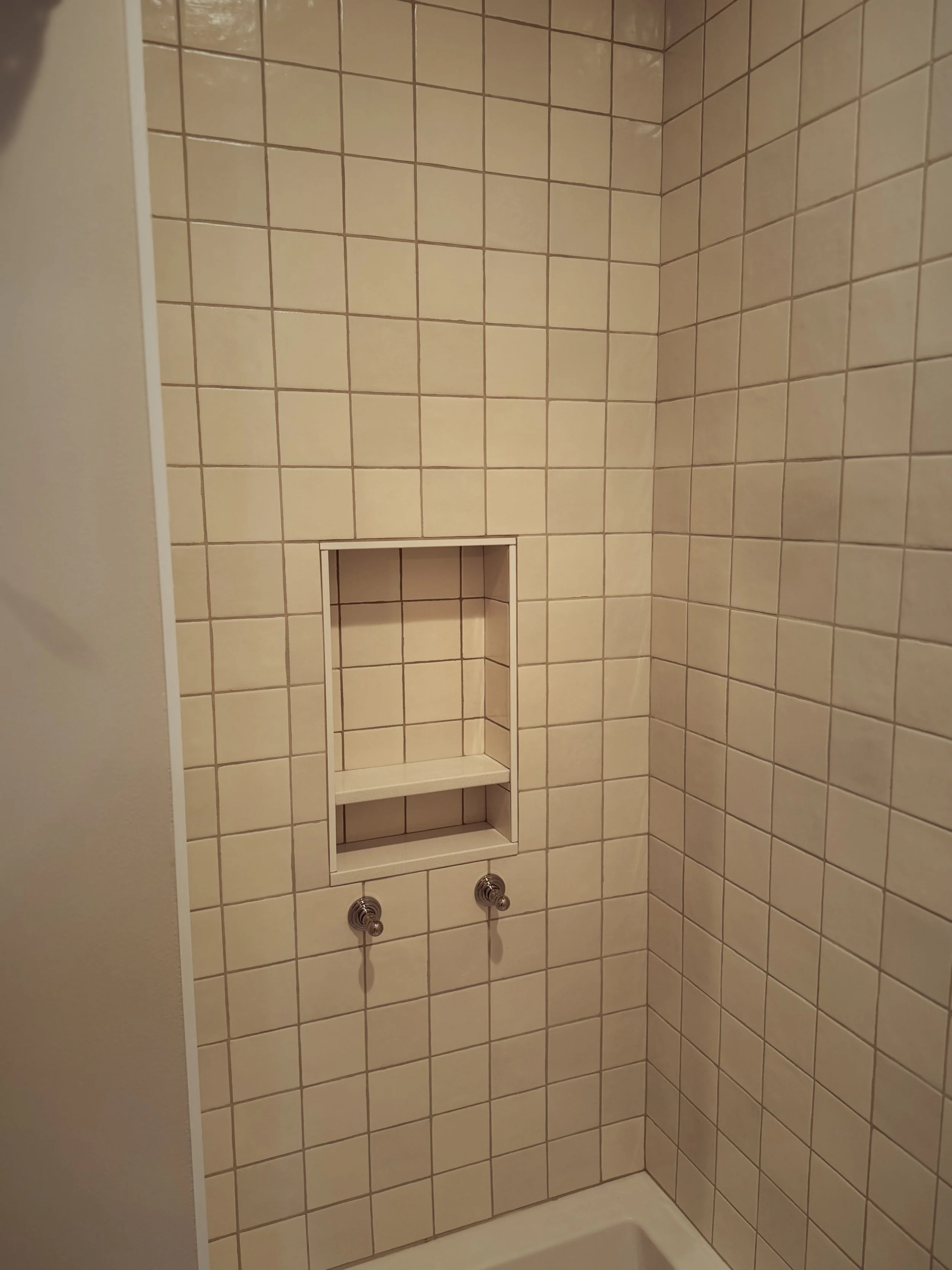 Bathroom shower wall with beige tiles, a built-in square shelf, and two shower knobs.