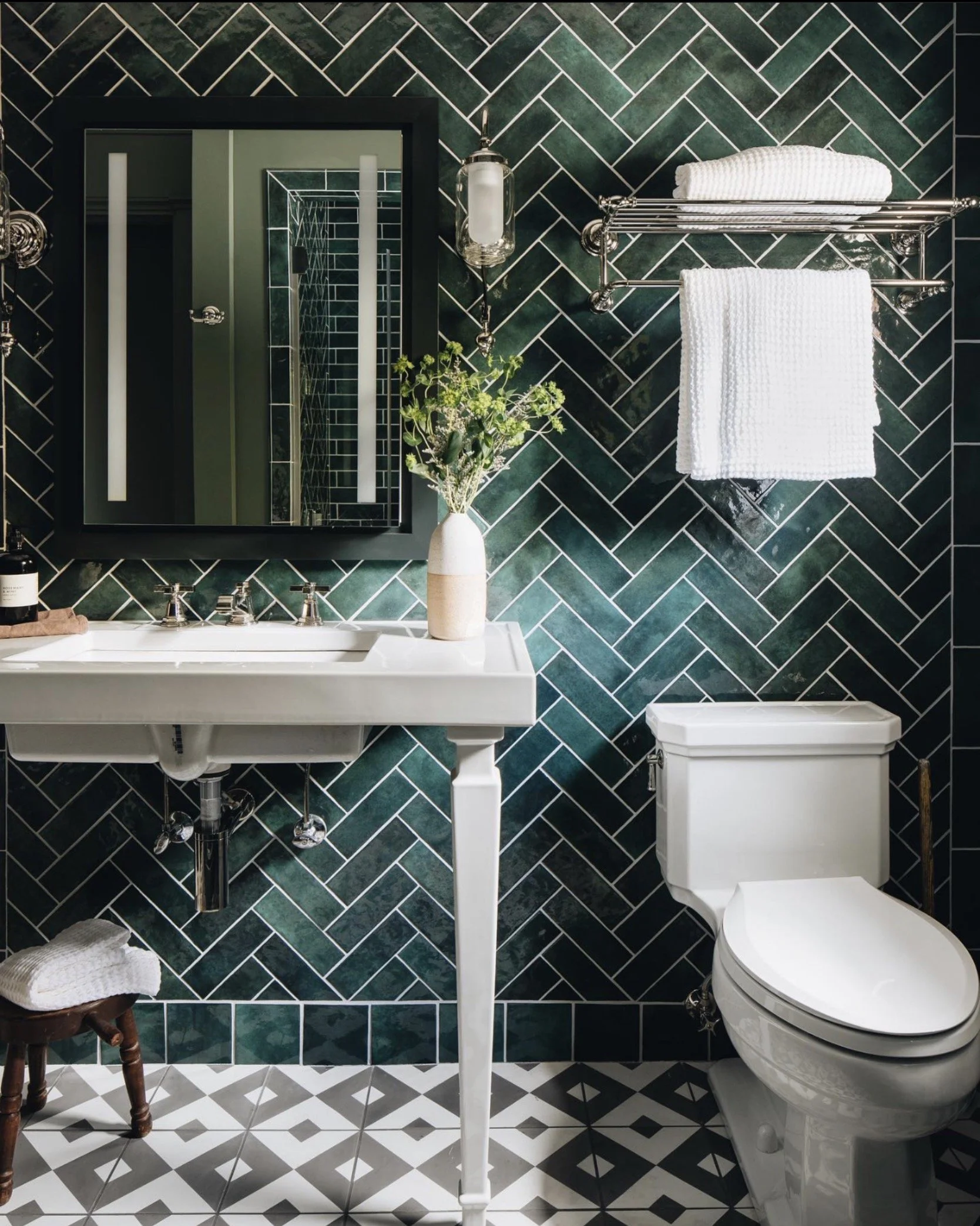 A modern bathroom featuring a white sink with a black-framed mirror above, green tile walls in a herringbone pattern, a white toilet, a metal shelf with towels, a vase with green plants, and a small wooden stool with a towel on it.