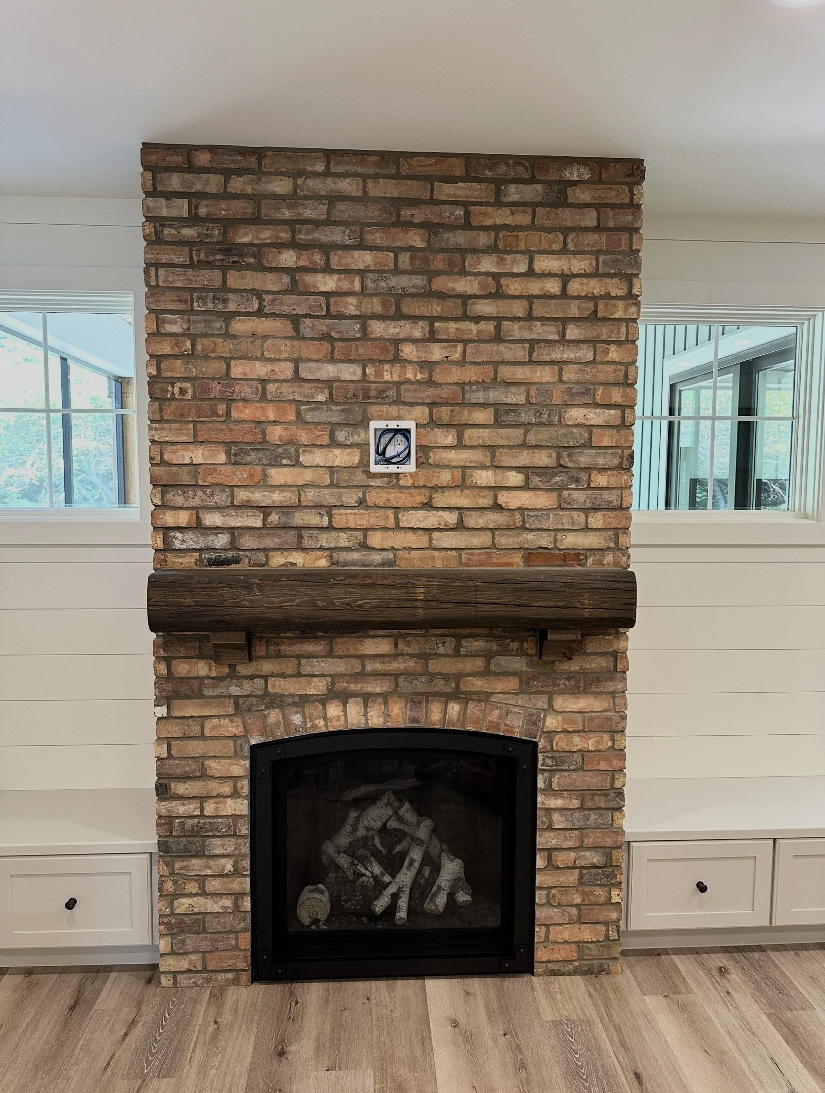 Interior brick fireplace with a wooden mantel and a black metal firebox, flanked by white wood-paneled walls and two windows.