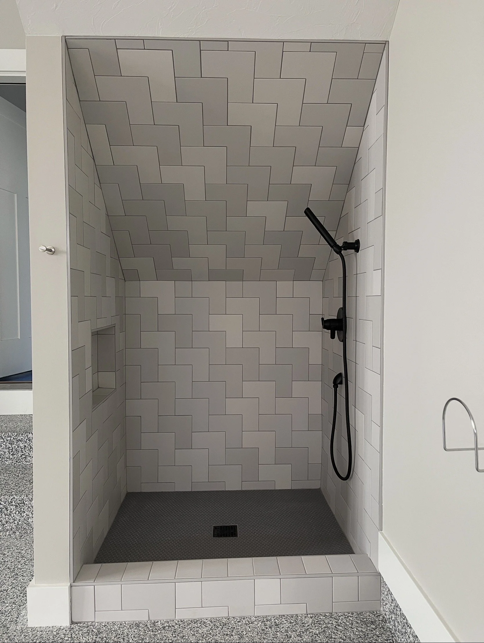 Modern walk-in shower with white subway tile walls, a black shower head and fixtures, and a gray textured floor with a drain in the center.