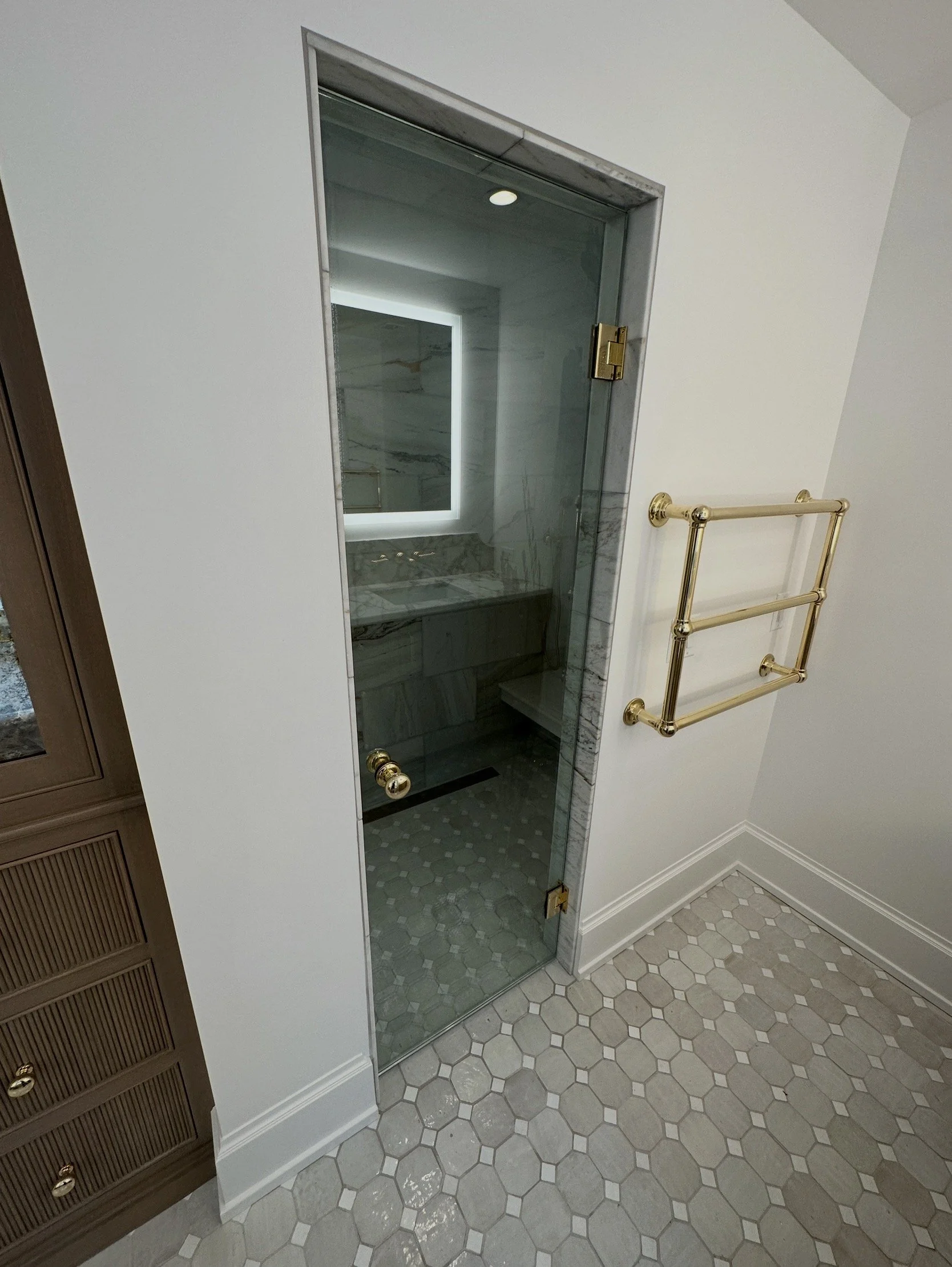 A walk-in shower with glass door, marble walls, and hexagonal floor tiles, adjacent to a white wall with a gold towel rack.