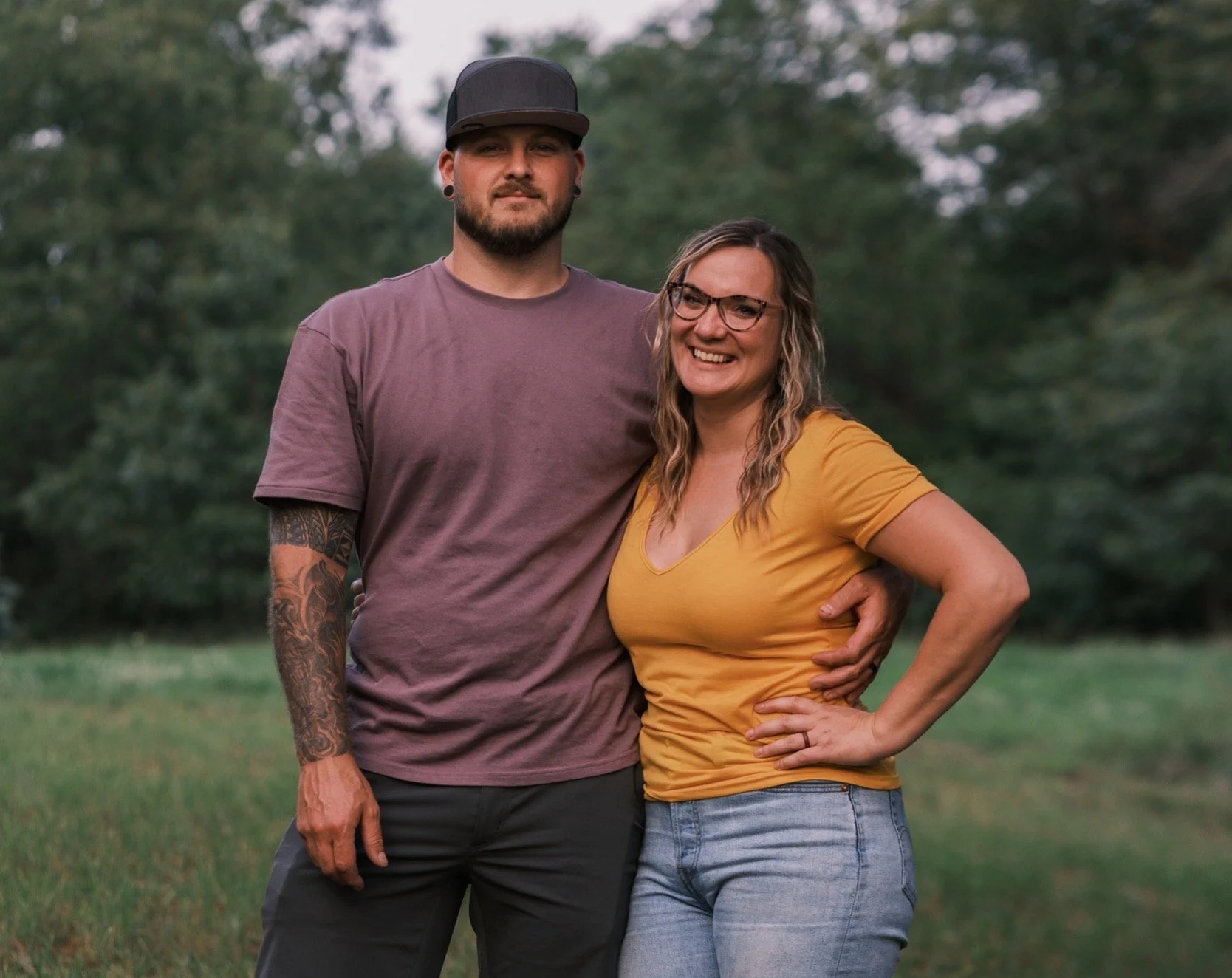A man and woman standing outdoors on a grassy area with trees in the background. The man, wearing a gray cap, purple t-shirt, and with tattoos on his left arm, has his arm around the woman. The woman, wearing glasses, a yellow top, and jeans, is smiling and has her hand on her hip.