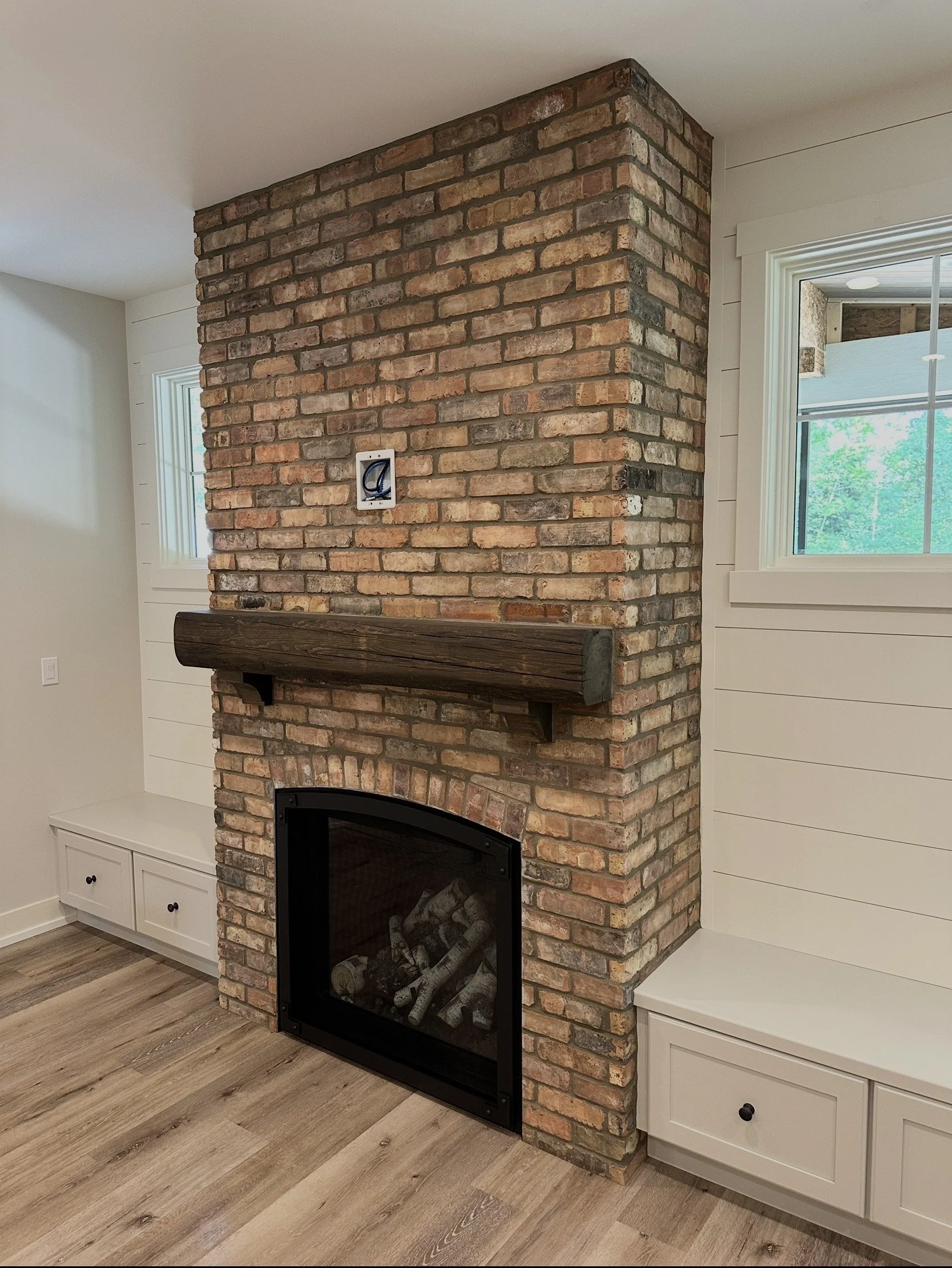 Interior view of a brick fireplace with a wooden mantel and a built-in storage bench with drawers on each side, in a room with wood flooring and white paneled walls.