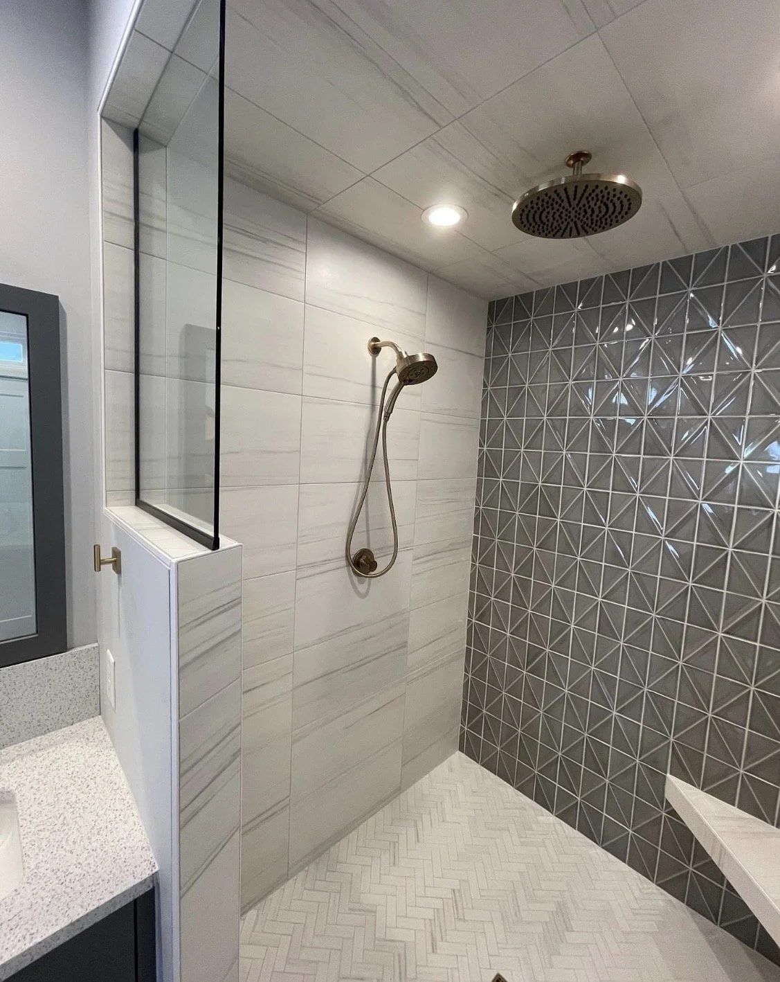 Modern bathroom shower area with patterned gray tiles on one wall, white tiles on another, a ceiling-mounted rainfall showerhead, and a handheld showerhead. A partial view of a countertop with a speckled surface is visible on the left.