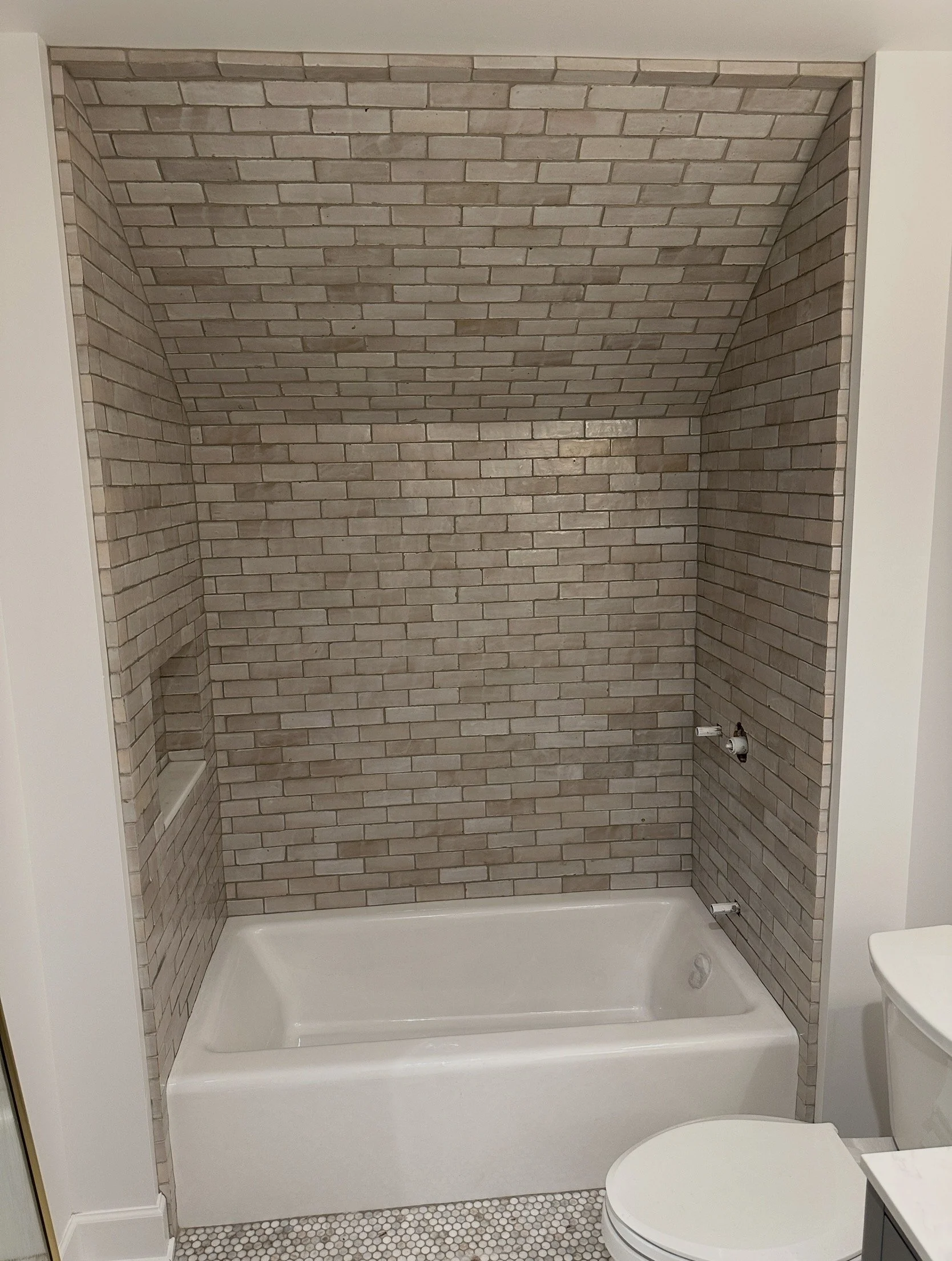 Bathroom with brick accent wall in shower area, white bathtub, and toilet, with small hexagonal floor tiles.