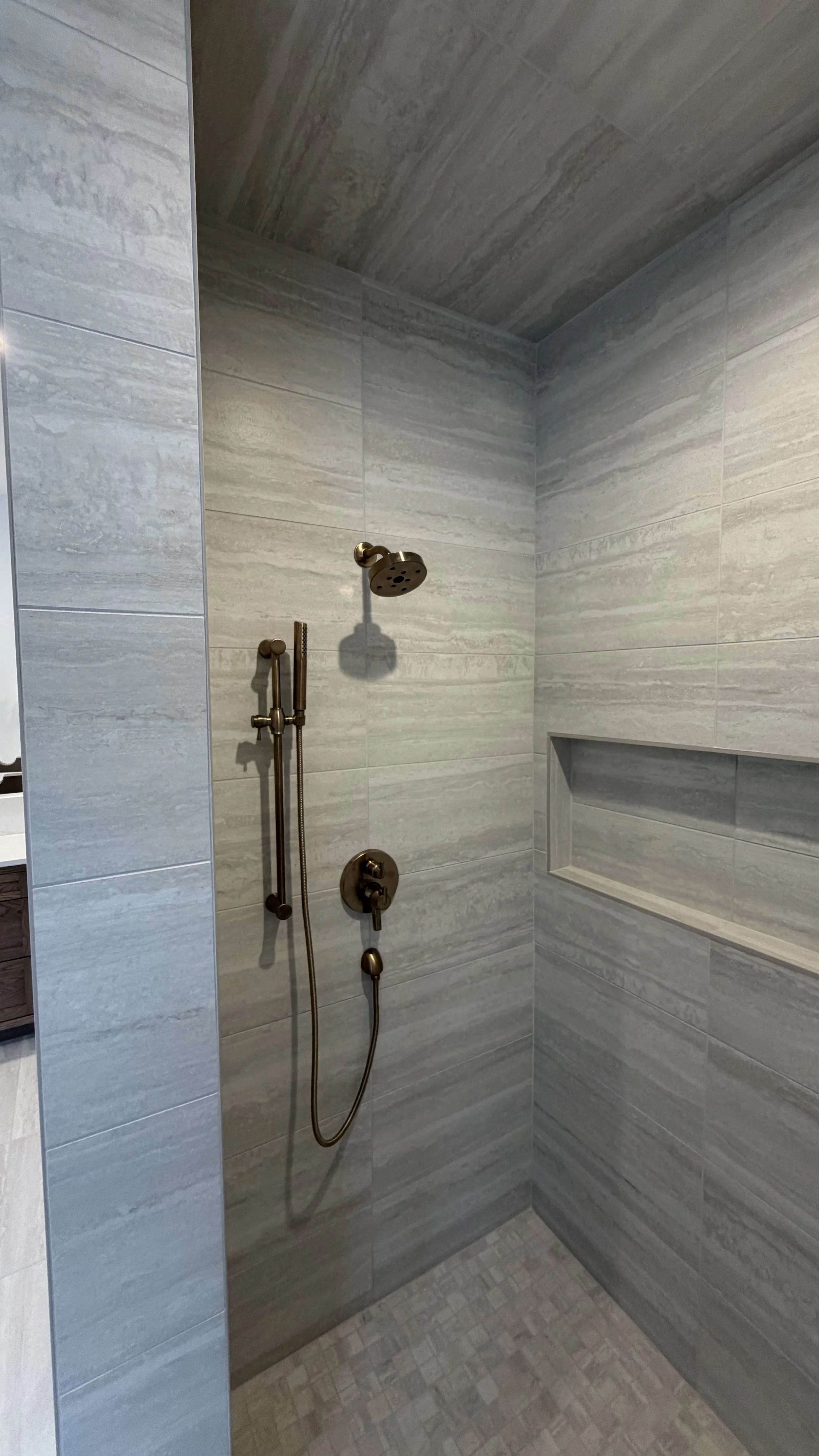 A walk-in shower with gray tiles, a brass shower head, a handheld shower wand, and a recessed shelf in the wall.