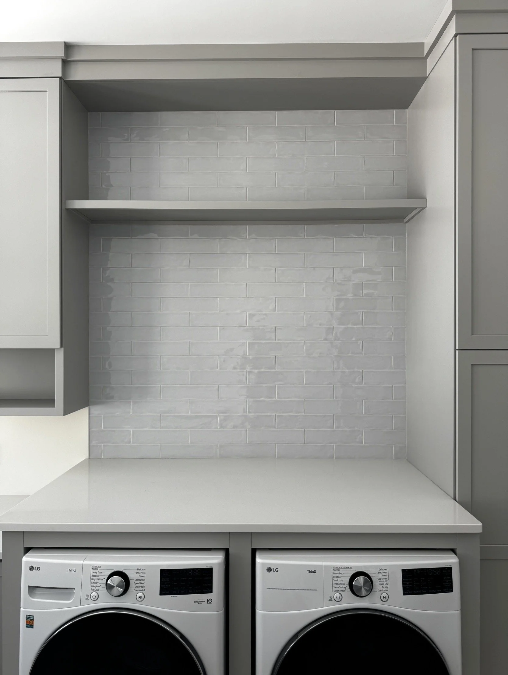 Laundry room with gray cabinets, a white countertop, a gray brick backsplash, and two front-loading washing machines.
