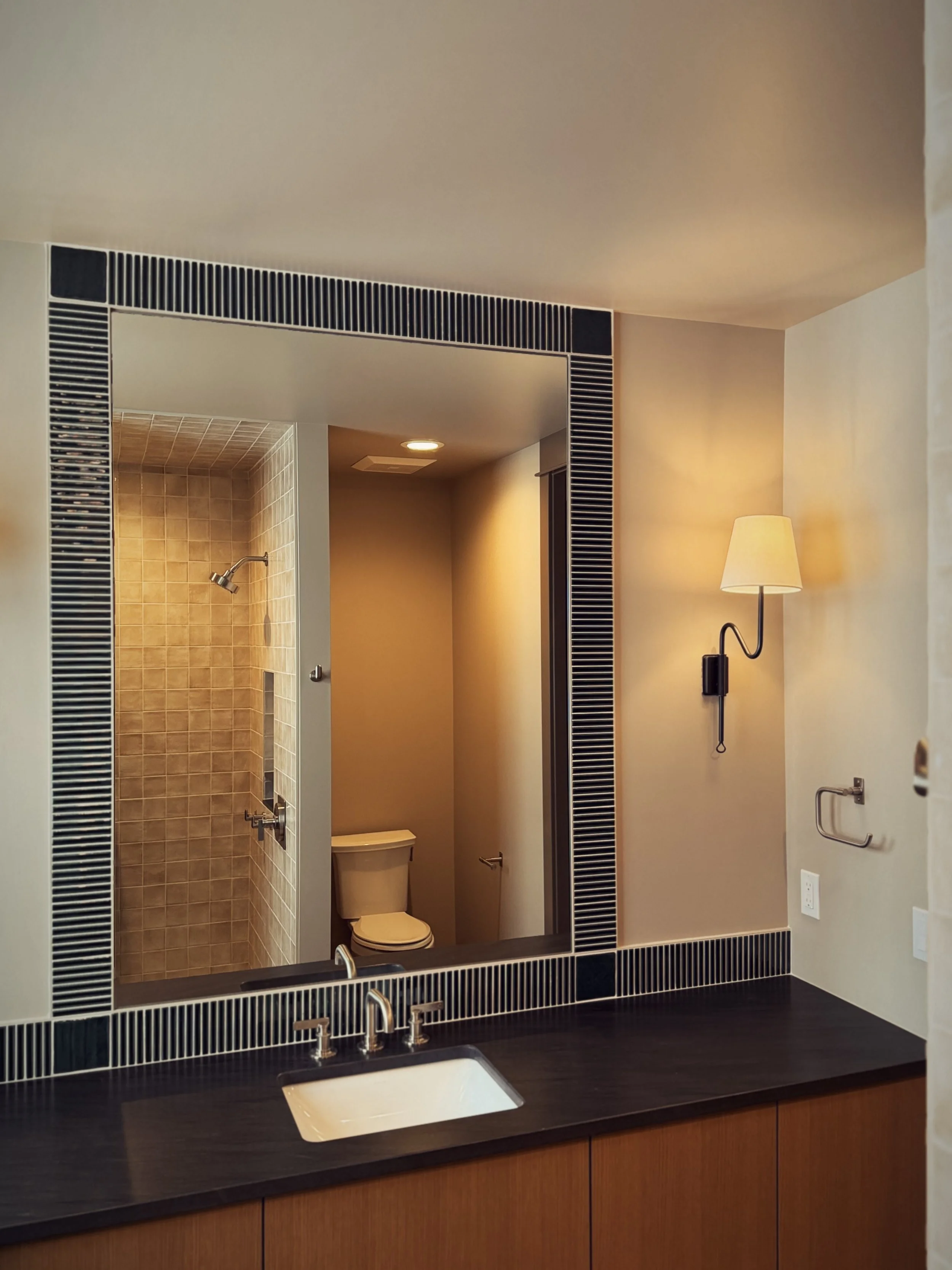Modern bathroom with a large mirror, black and white tile border, black countertop, white sink, wall-mounted lamp, and shower and toilet visible through reflection.