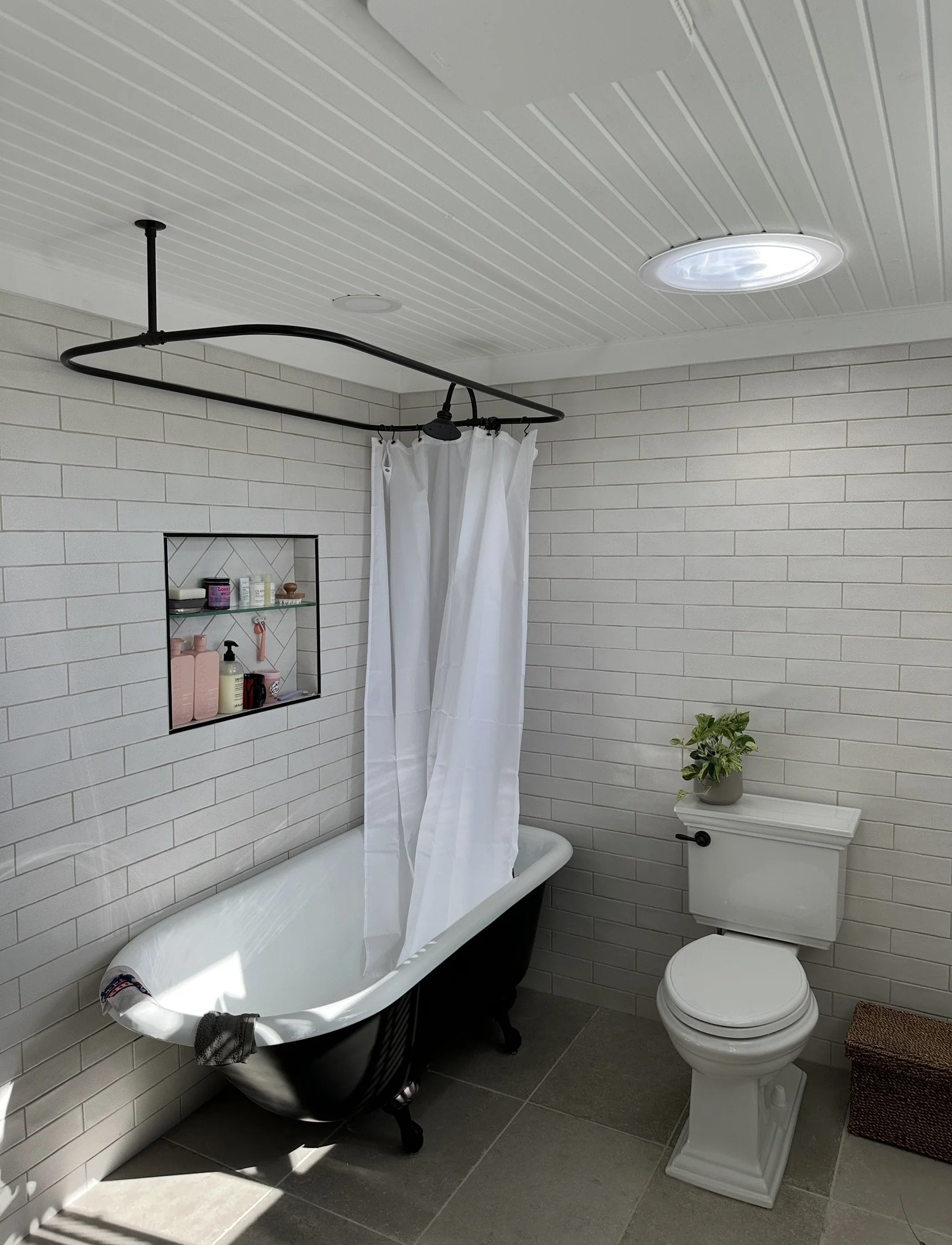 Bathroom with clawfoot bathtub, black exterior, white interior, white shower curtain, shelf with toiletries, white brick walls, ceiling with round light, potted plant on toilet tank, gray tile floor, woven brown storage basket.