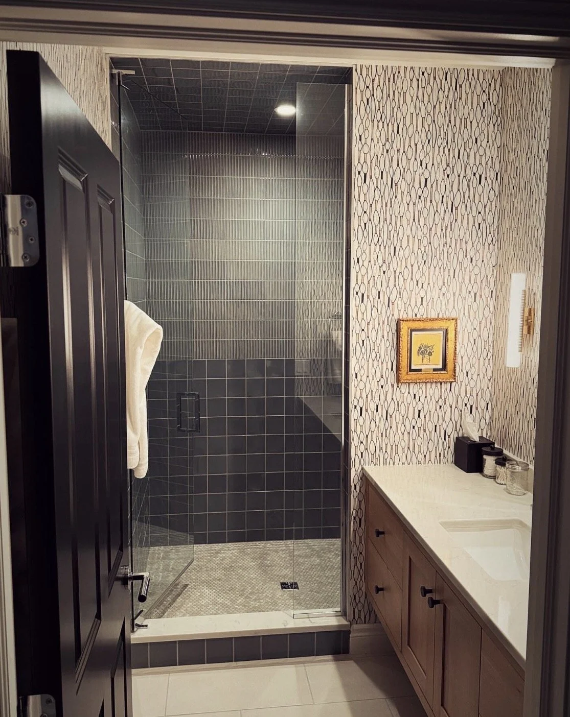 Modern bathroom with a walk-in shower enclosed by glass, a wooden vanity with a white countertop and a sink, a patterned wallpaper, a framed picture on the wall, and a tissue box with jars on the countertop.