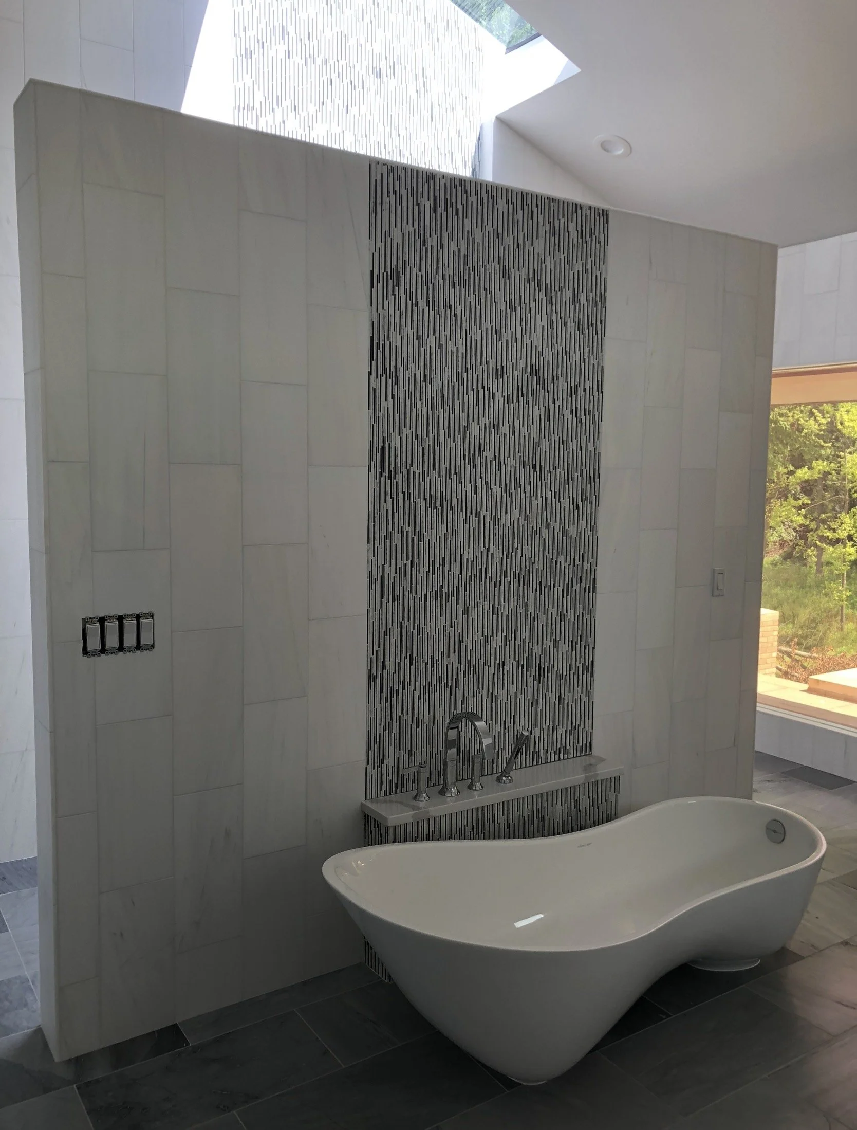 Modern bathroom with a white freestanding bathtub, gray tiled floor, a decorative black and white mosaic wall, and a window showing greenery outside.