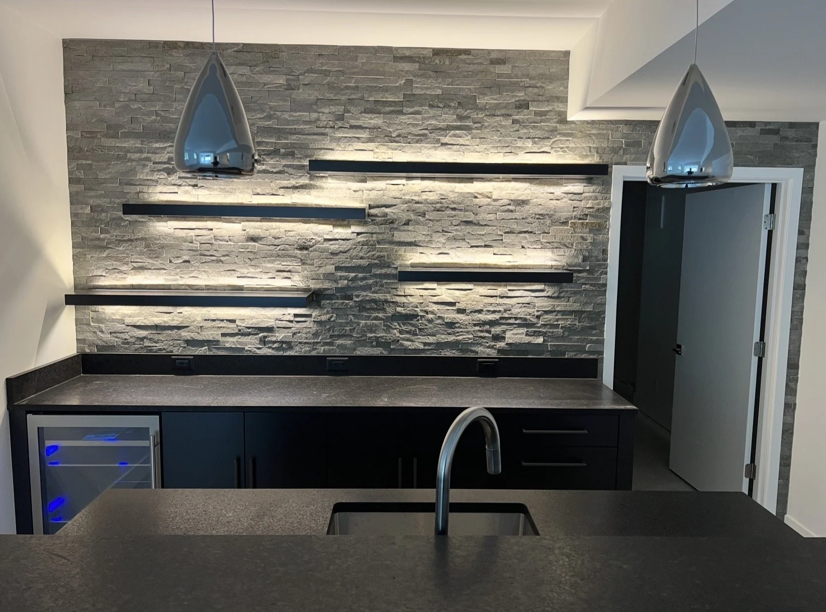 Modern kitchen with black cabinetry, a stone accent wall, floating shelves, a minimalistic sink, and pendant lights.