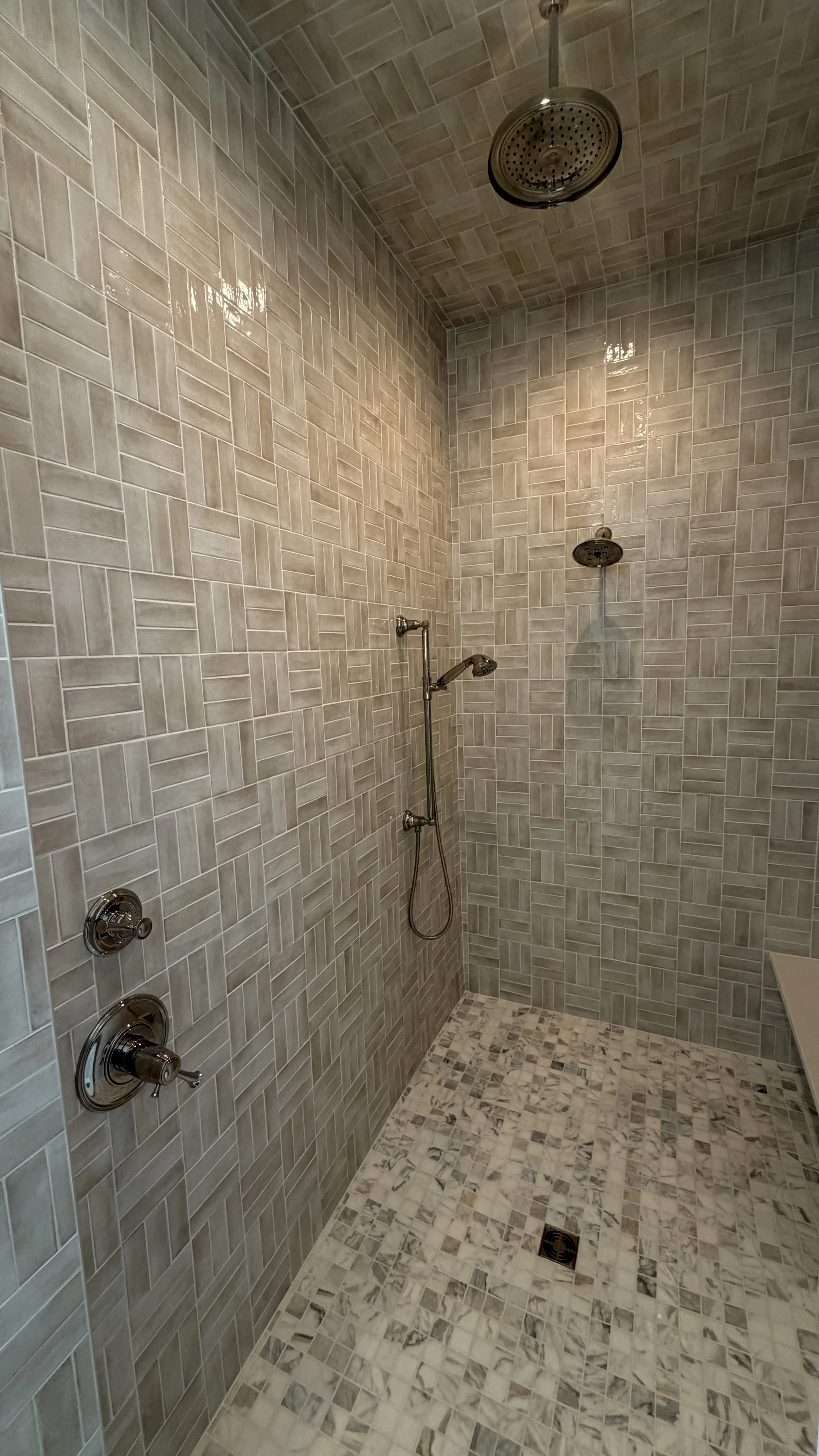 Bathroom shower with beige and gray tile walls and floor, two showerheads, and a wall-mounted control panel.