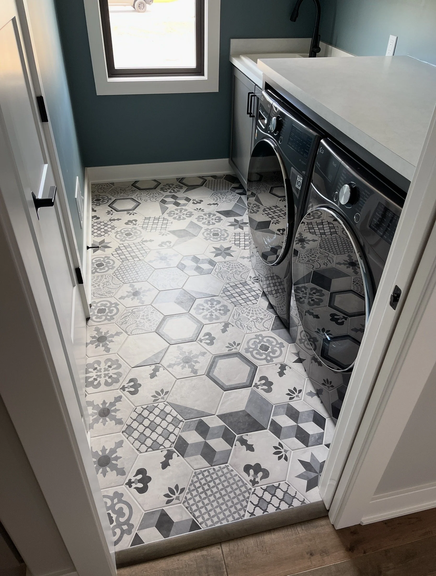 Laundry room with patterned tile floor, blue walls, white cabinets, a window, and a front-loading washing machine and dryer.