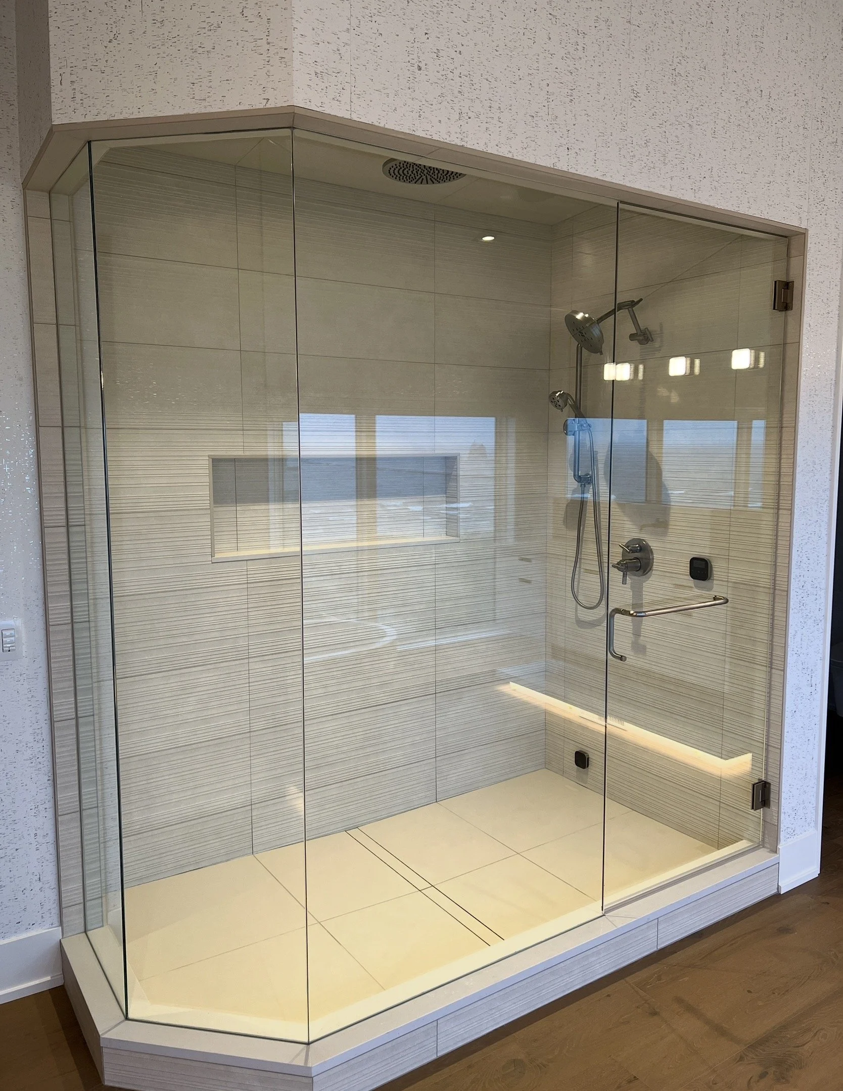 A modern corner shower with glass doors, beige tiled walls, built-in niche, and multiple shower heads, including a hand-held and a rainfall shower.