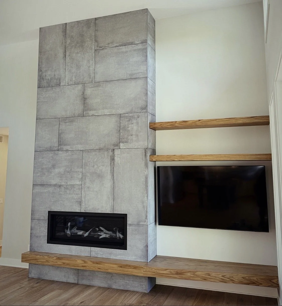 Living room corner with gray tiled fireplace, two wooden shelves, and a mounted flat-screen TV.