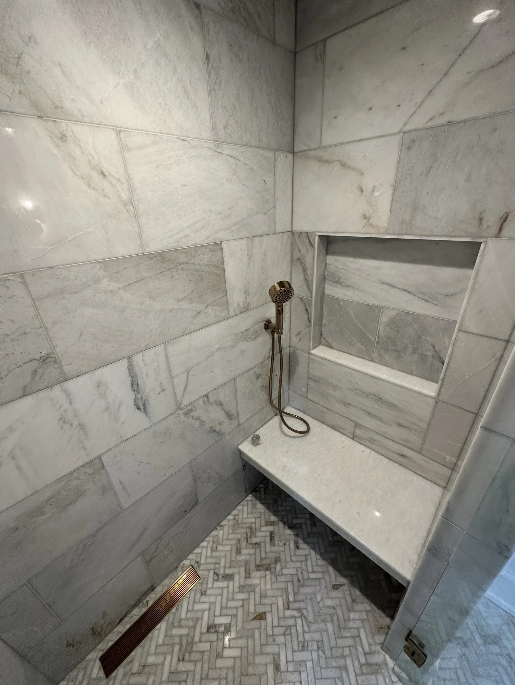 Marble tile shower with a brass handheld shower head, built-in shelf, and herringbone marble floor.