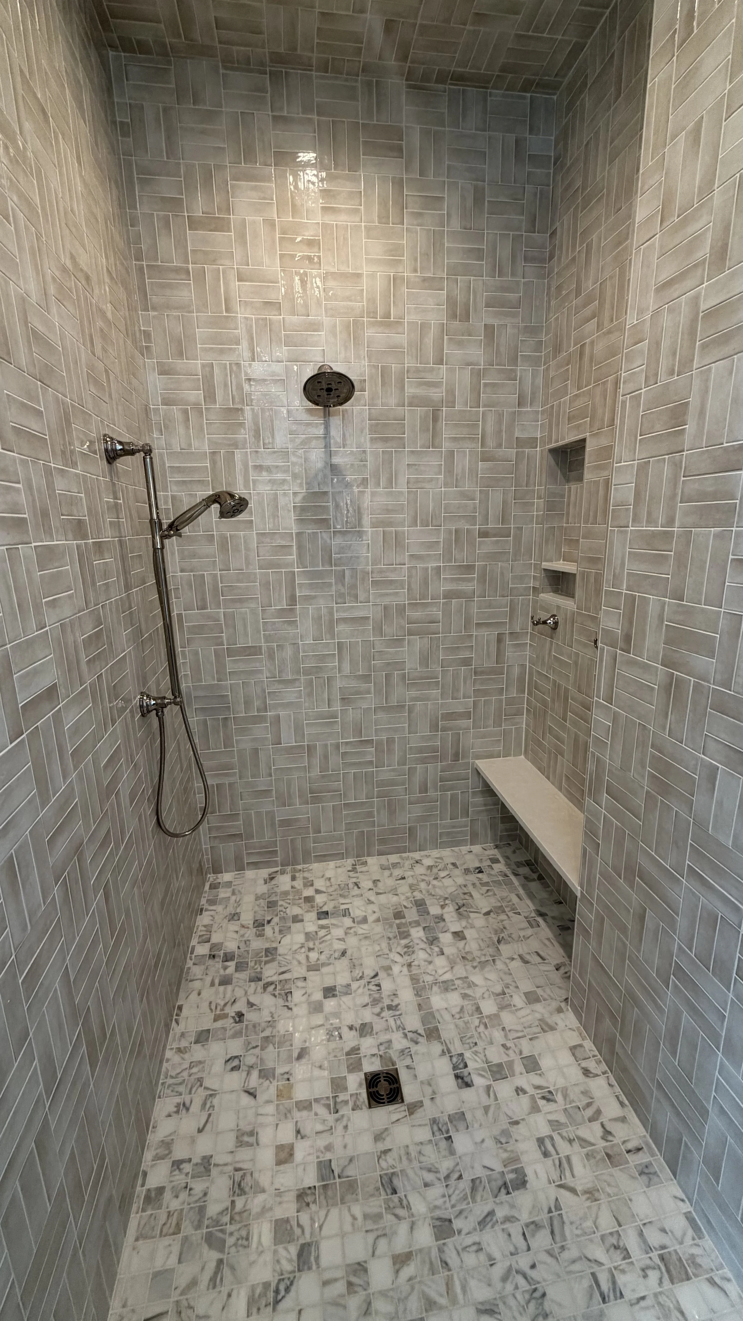 Empty walk-in shower with beige and gray tiled walls, marble tiled floor, a rain showerhead, a handheld showerhead, wall niche shelves, a small bench, and a drain on the floor.