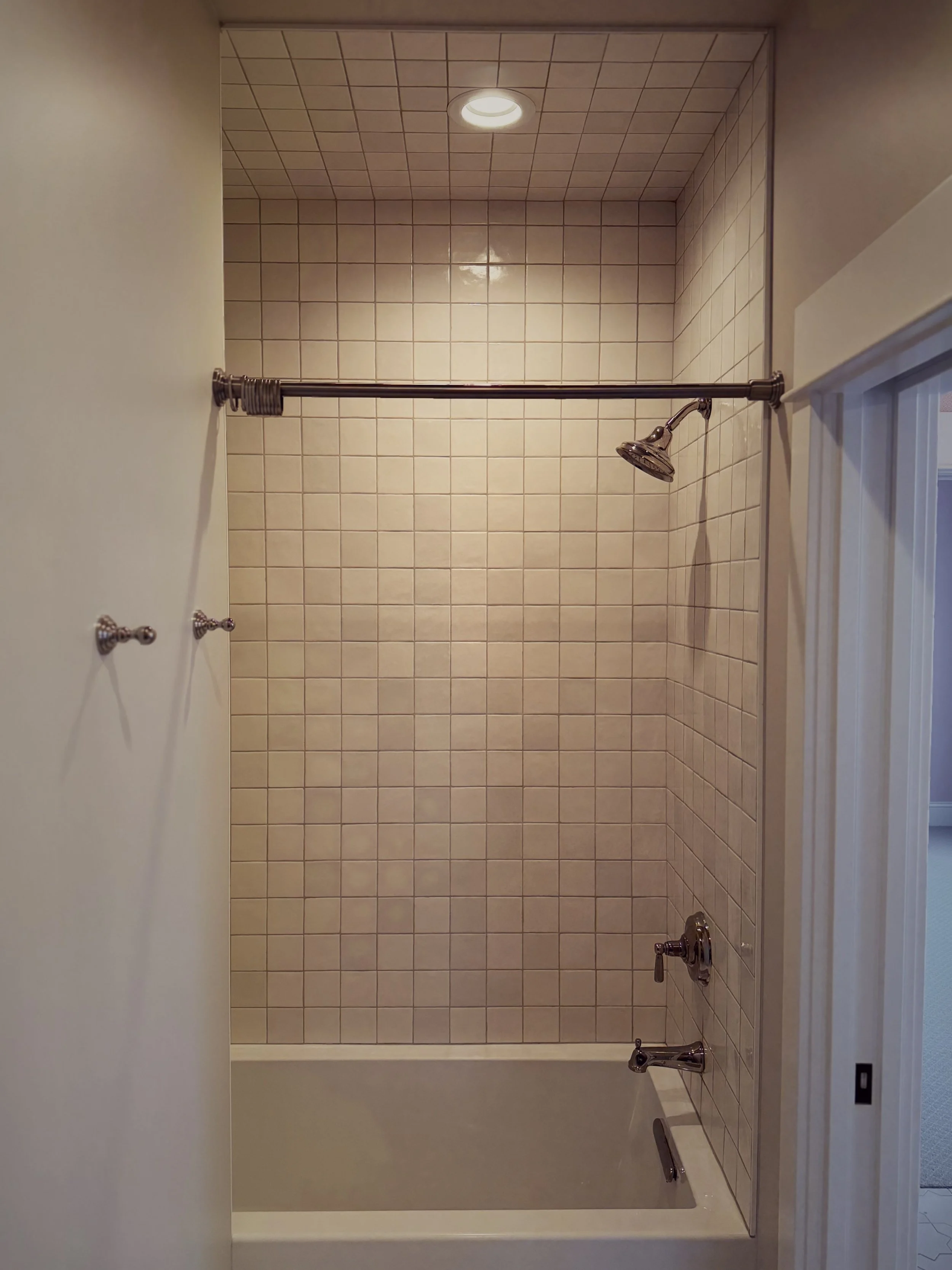 A bathroom shower area with beige tile walls, a showerhead on the right, three metal fixtures below it, and a ceiling light.