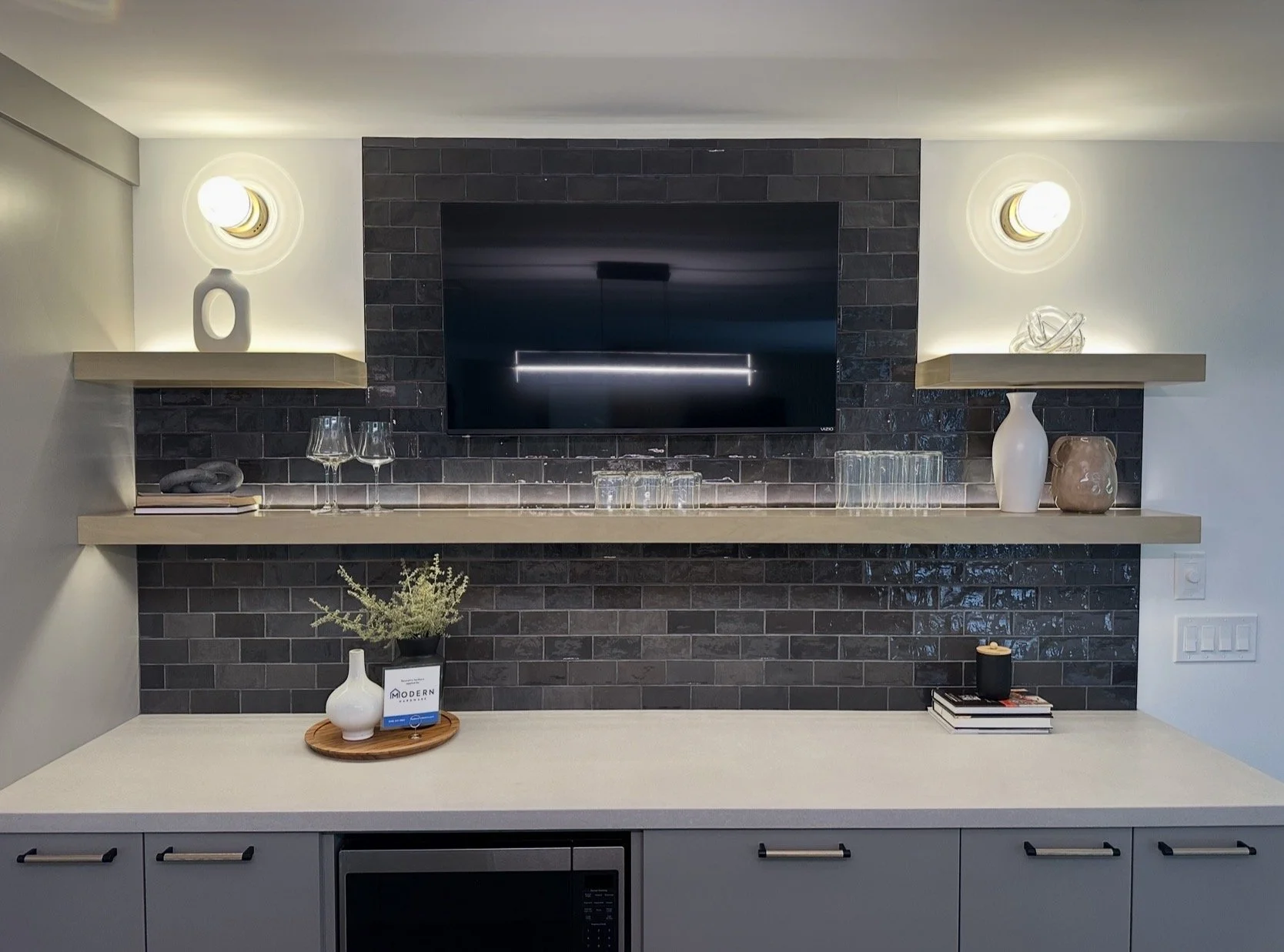 Modern kitchen with black tiled wall, mounted TV, floating shelves with decorative objects, glassware, and a built-in microwave, with a beige countertop and drawers.