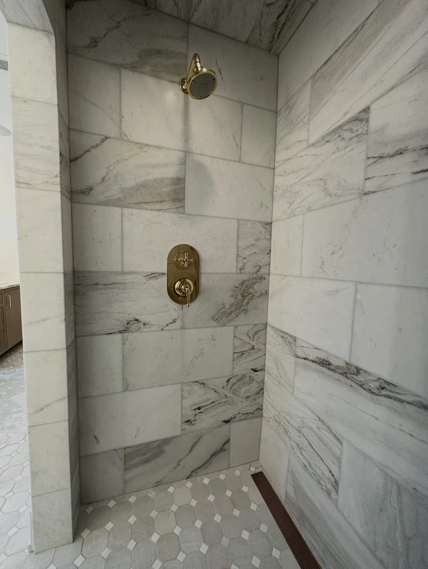 Shower area with marble tiles, a wall-mounted gold showerhead, and a gold control valve. The floor has beige hexagonal and small square tiles, with a linear floor drain along the edge.