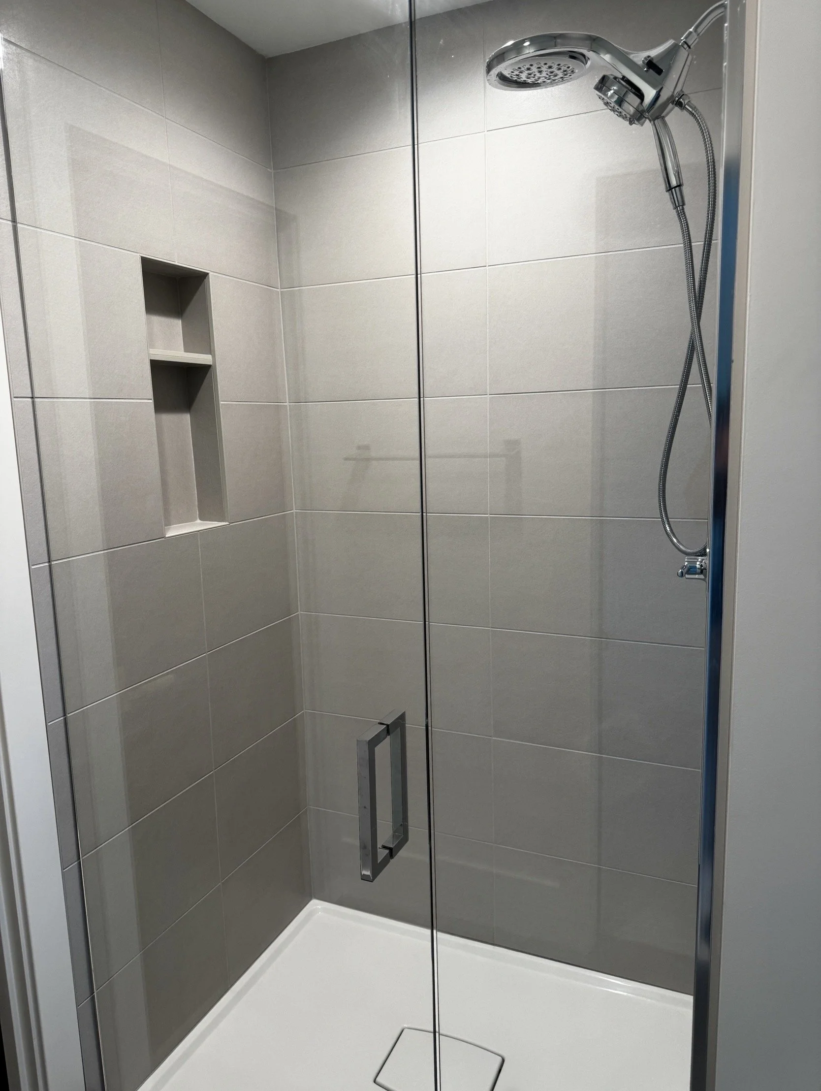 Empty walk-in shower with beige tiles, a recessed shelf, a glass door with metal handle, a ceiling-mounted showerhead, and a white shower pan.