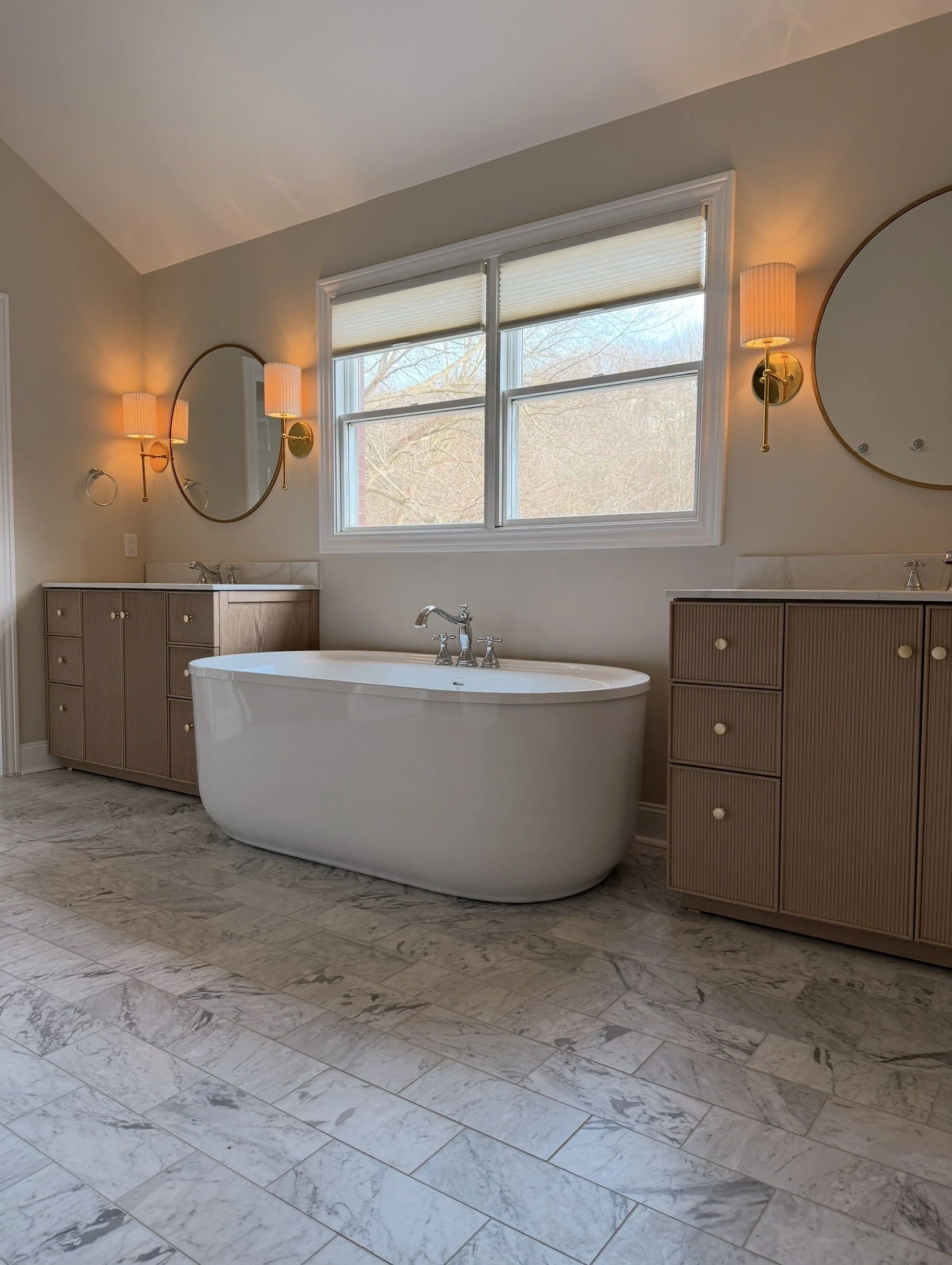 Modern bathroom featuring a freestanding bathtub, two wooden vanity cabinets with round gold knobs, round wall mirrors, wall sconces with gold fixtures, and a window with a view of leafless trees outside.