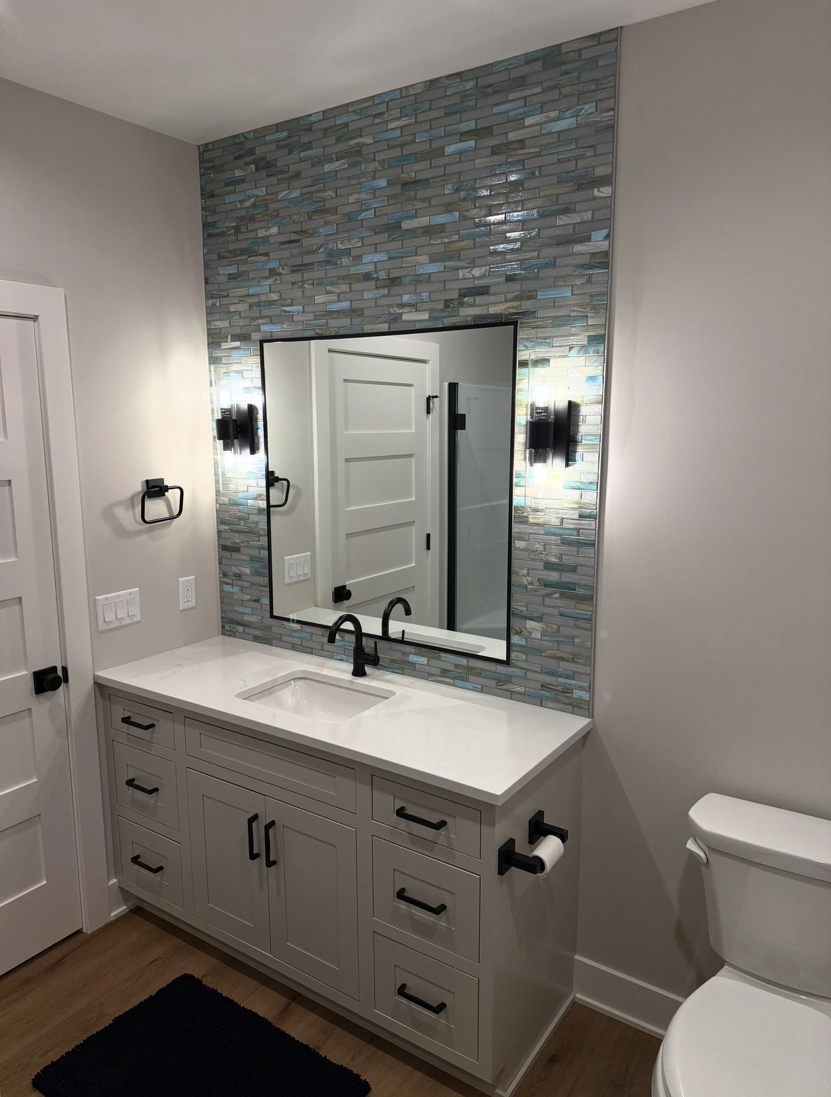 Modern bathroom with white vanity, black fixtures, large mirror, textured tile wall, wall-mounted sconces, toilet, and wood flooring.