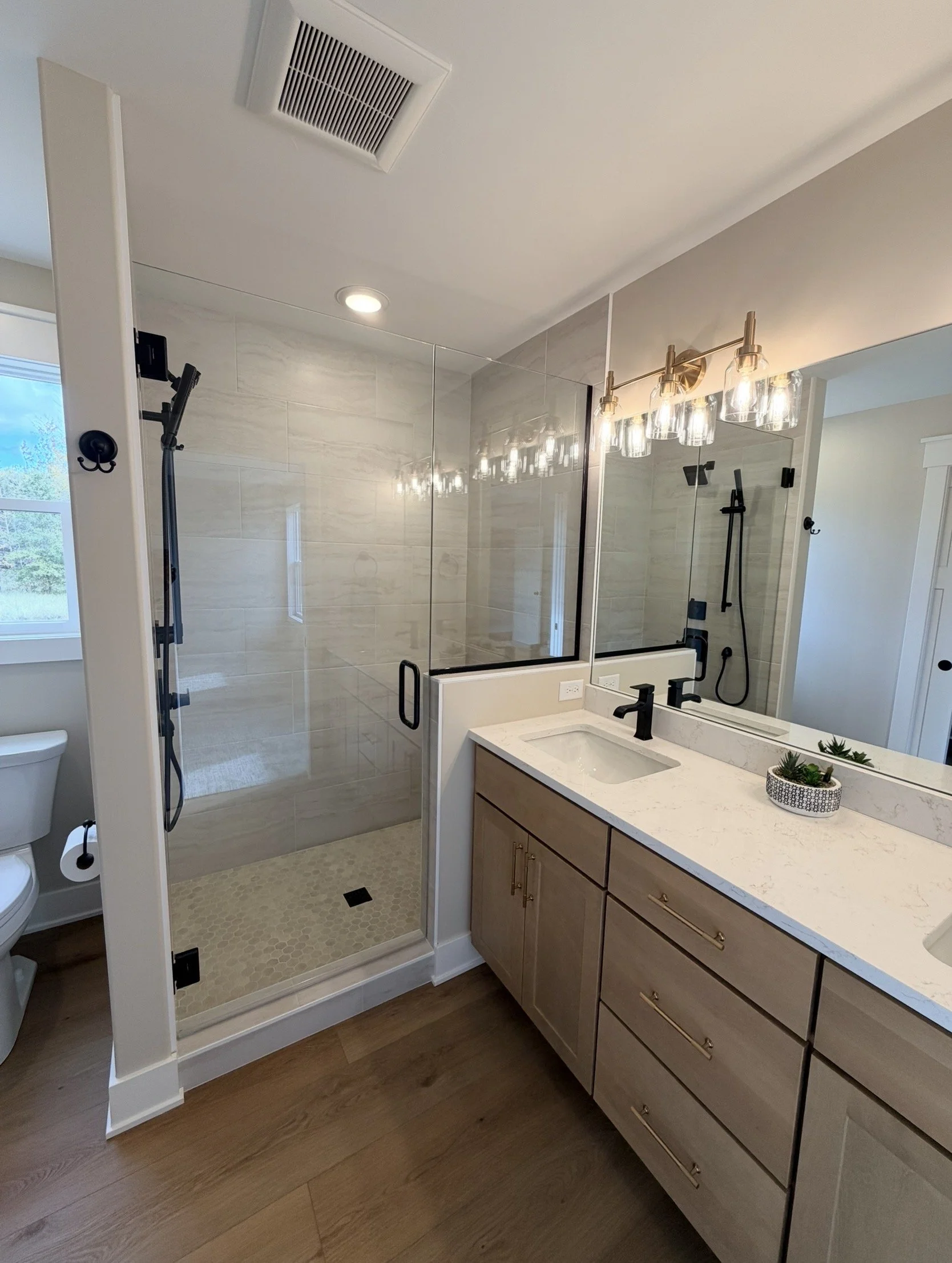 Modern bathroom with walk-in glass shower, white marble vanity countertop, wooden cabinet, large mirror, decorative plant, and soft lighting.