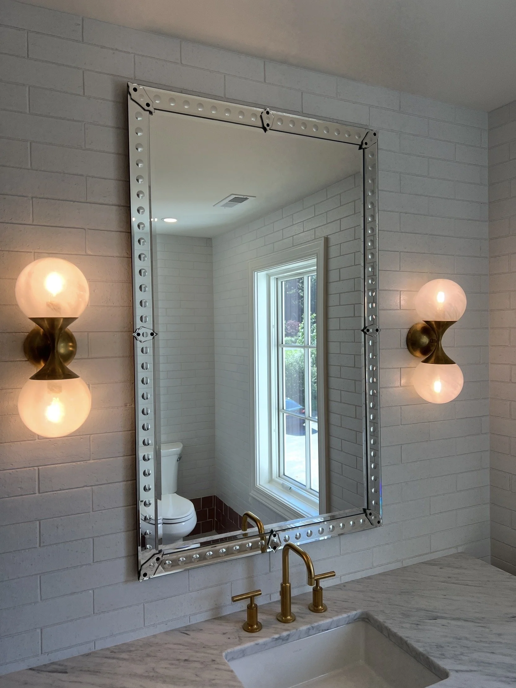 Bathroom with a large mirror, two sconces on either side, white brick walls, a window, and a marble countertop with a gold faucet.