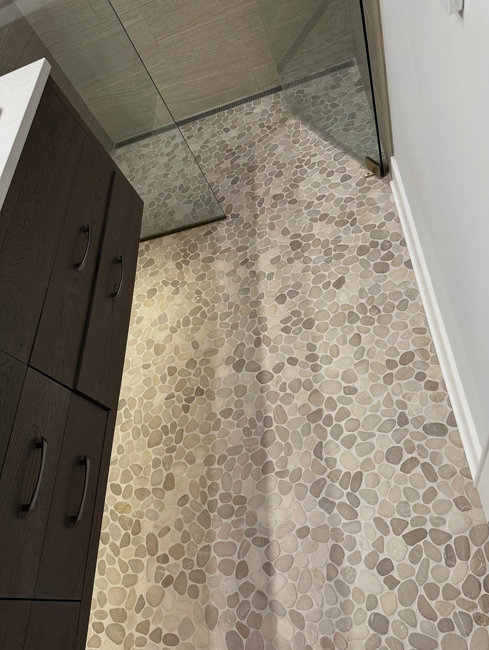 View of a bathroom floor with pebble tile pattern, part of a wooden cabinet on the left, a glass shower door at the top, and a white wall with trim on the right.