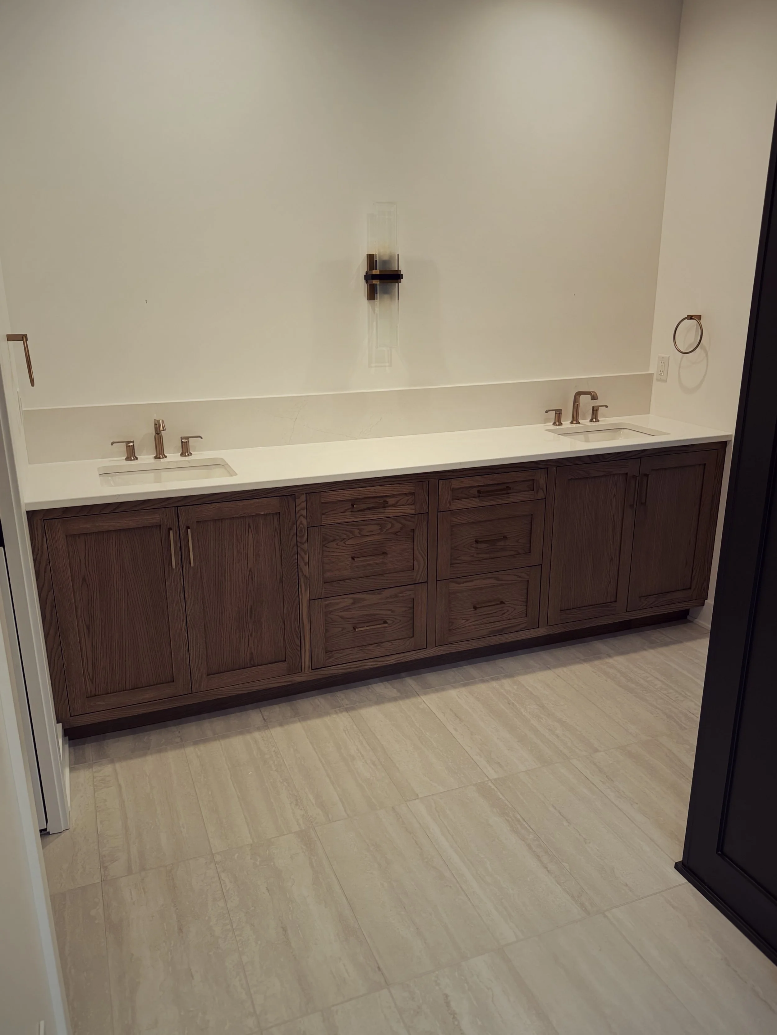 Bathroom vanity with double sinks, wooden cabinets, beige tile floor, and a wall-mounted light fixture