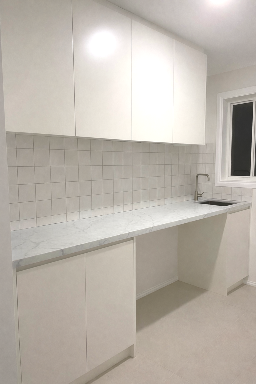 A minimalist kitchen with white cabinets, a white marble countertop, a tiled backsplash, a small sink with a modern faucet, and a window with a white frame.