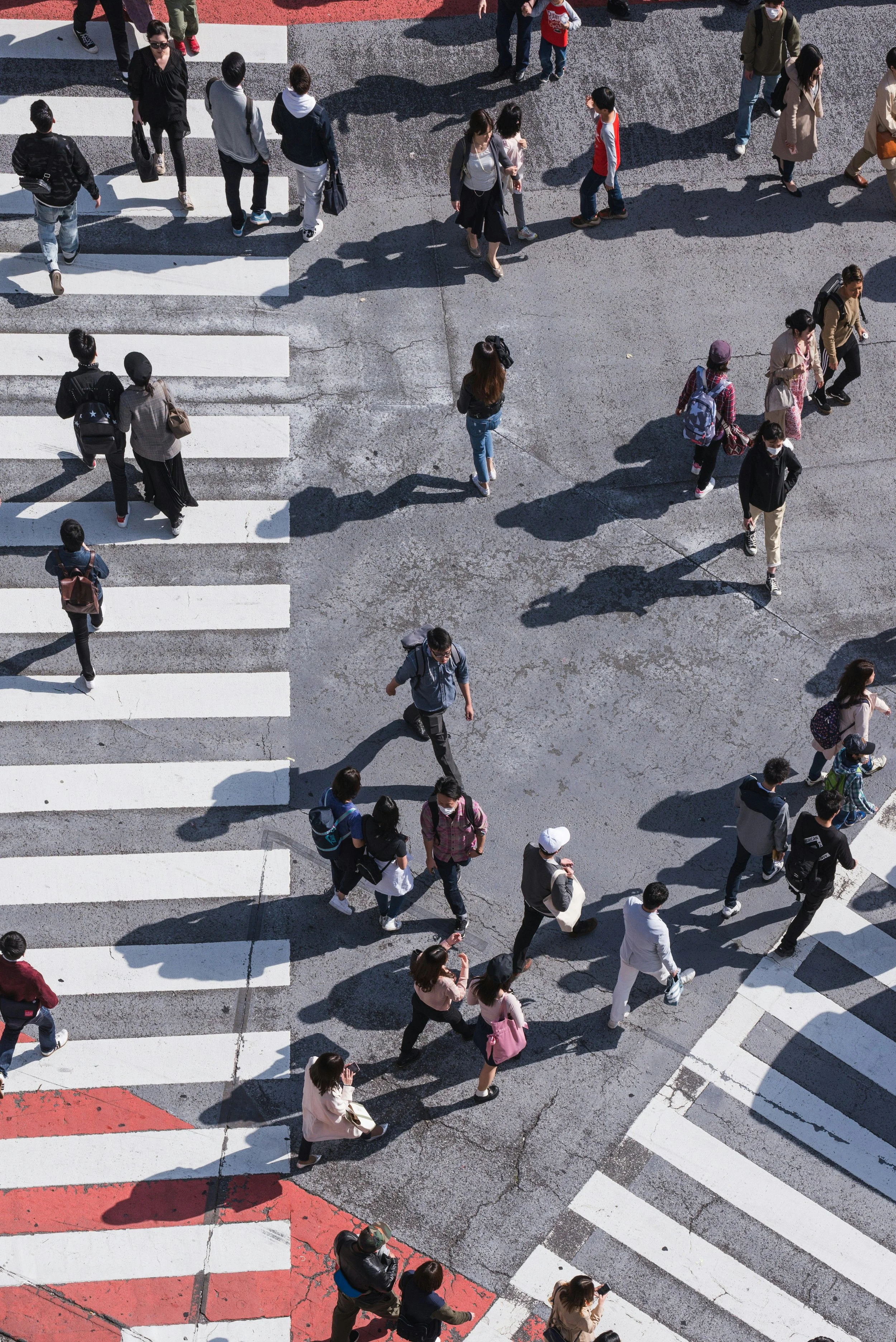 Aerial view of a busy city crosswalk with pedestrians walking in various directions. The crosswalk has white stripes on gray pavement, with some red sections at the edges. People are wearing casual clothing and some are carrying backpacks and umbrellas.