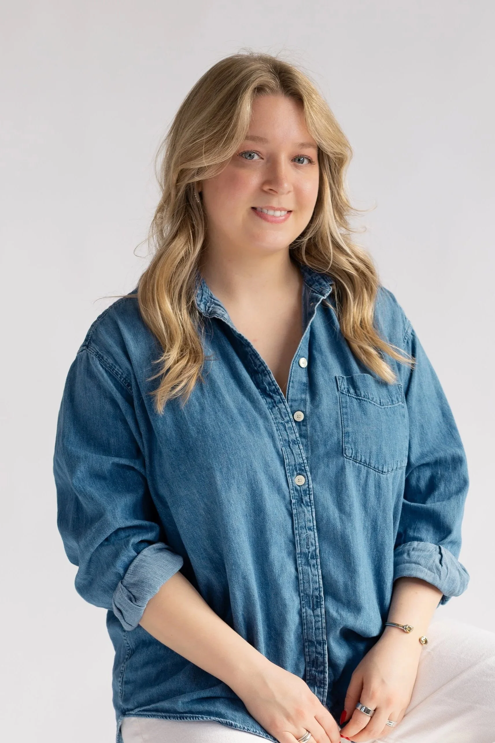 A young woman with blonde hair, wearing a denim shirt, smiling at the camera against a plain light gray background.