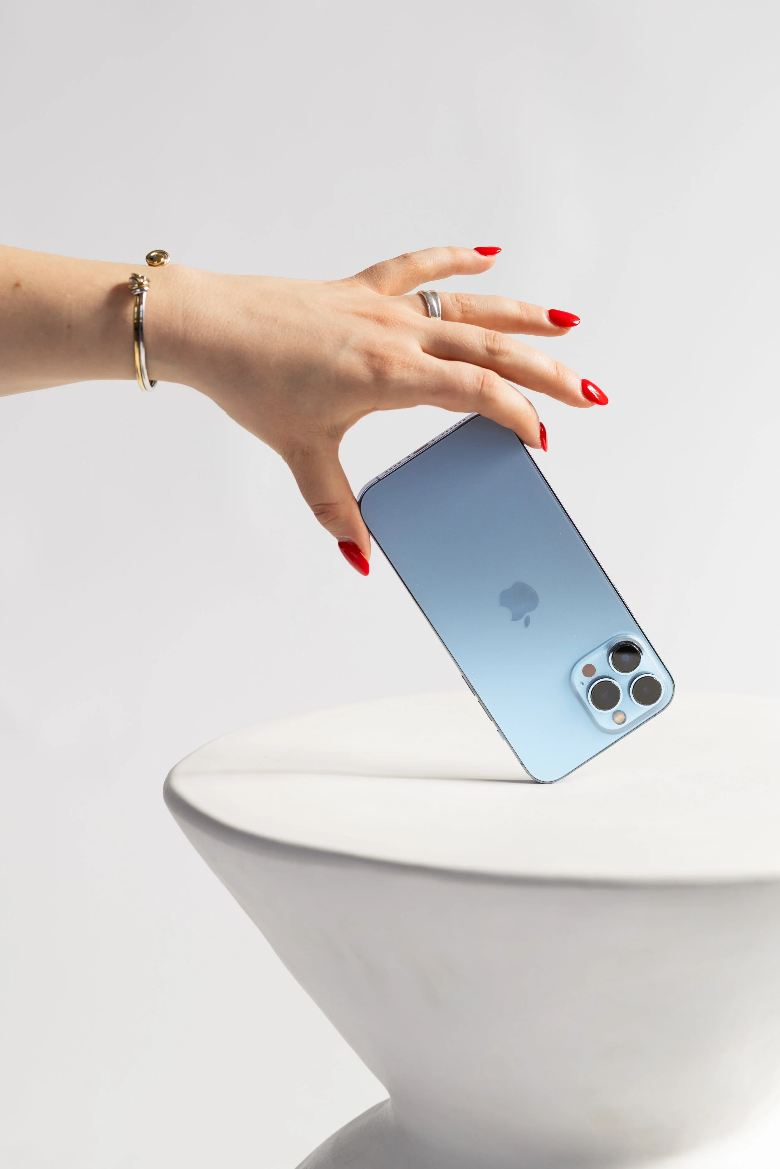 A hand with red-painted nails and silver jewelry is holding a blue iPhone 13 Pro, with the Apple logo visible, over a white, modern, curved table against a plain white background.