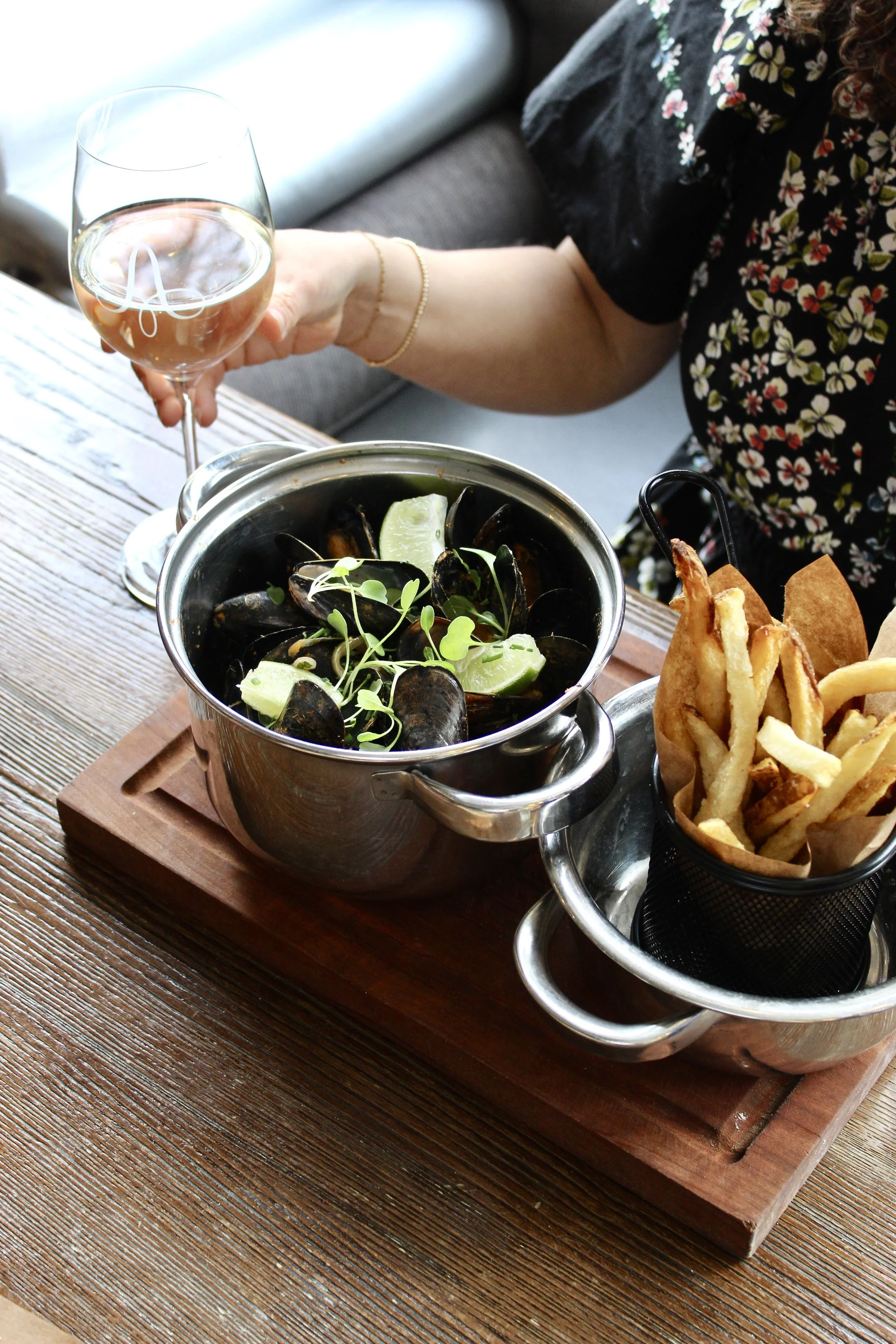 Table setting with a bowl of mussels garnished with lime wedges and microgreens, a side of French fries, a glass of white wine, and a woman in floral dress holding the wine glass.