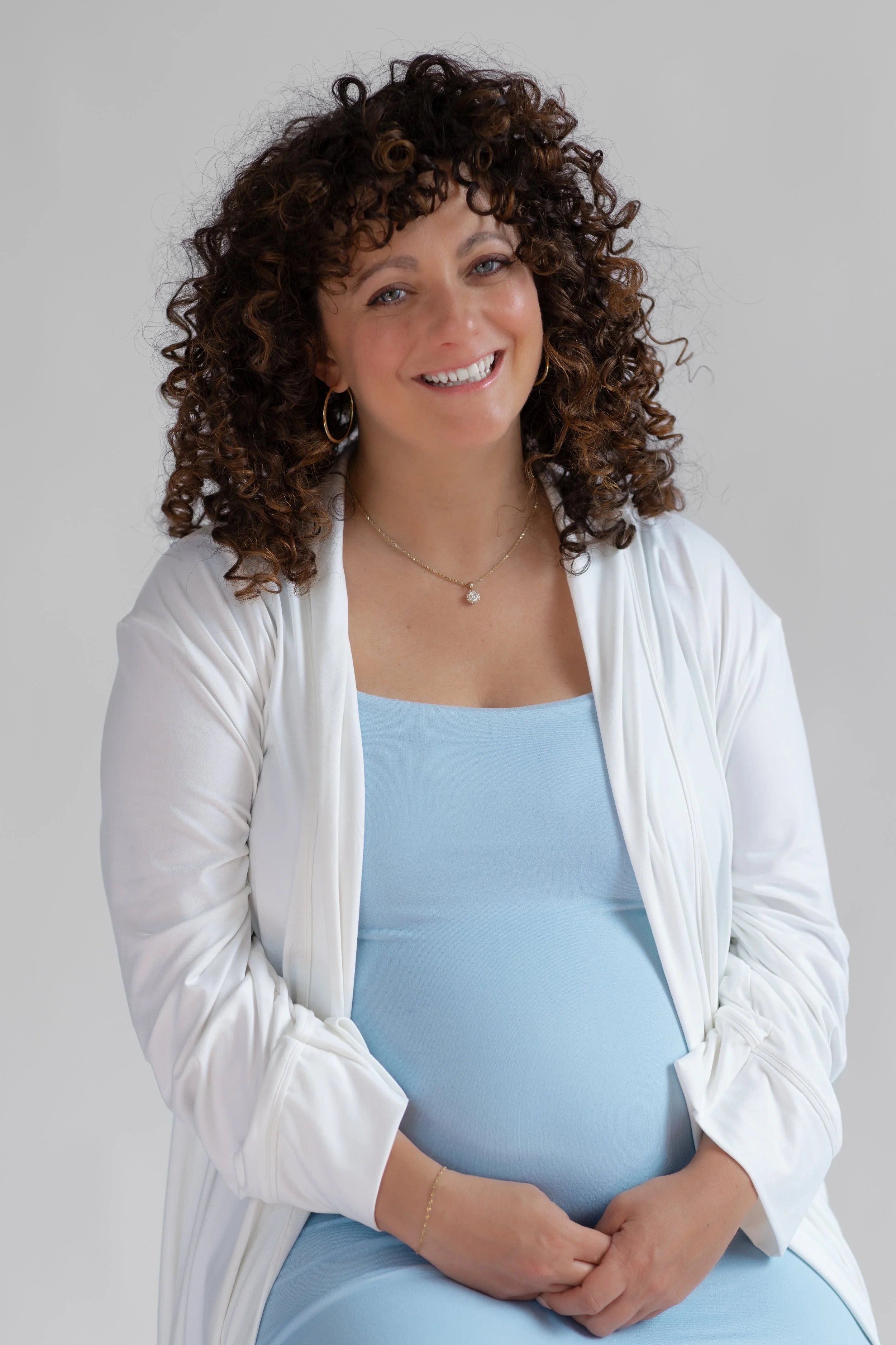Woman with curly brown hair wearing a light blue dress, white cardigan, gold jewelry, smiling, sitting against a light gray background.