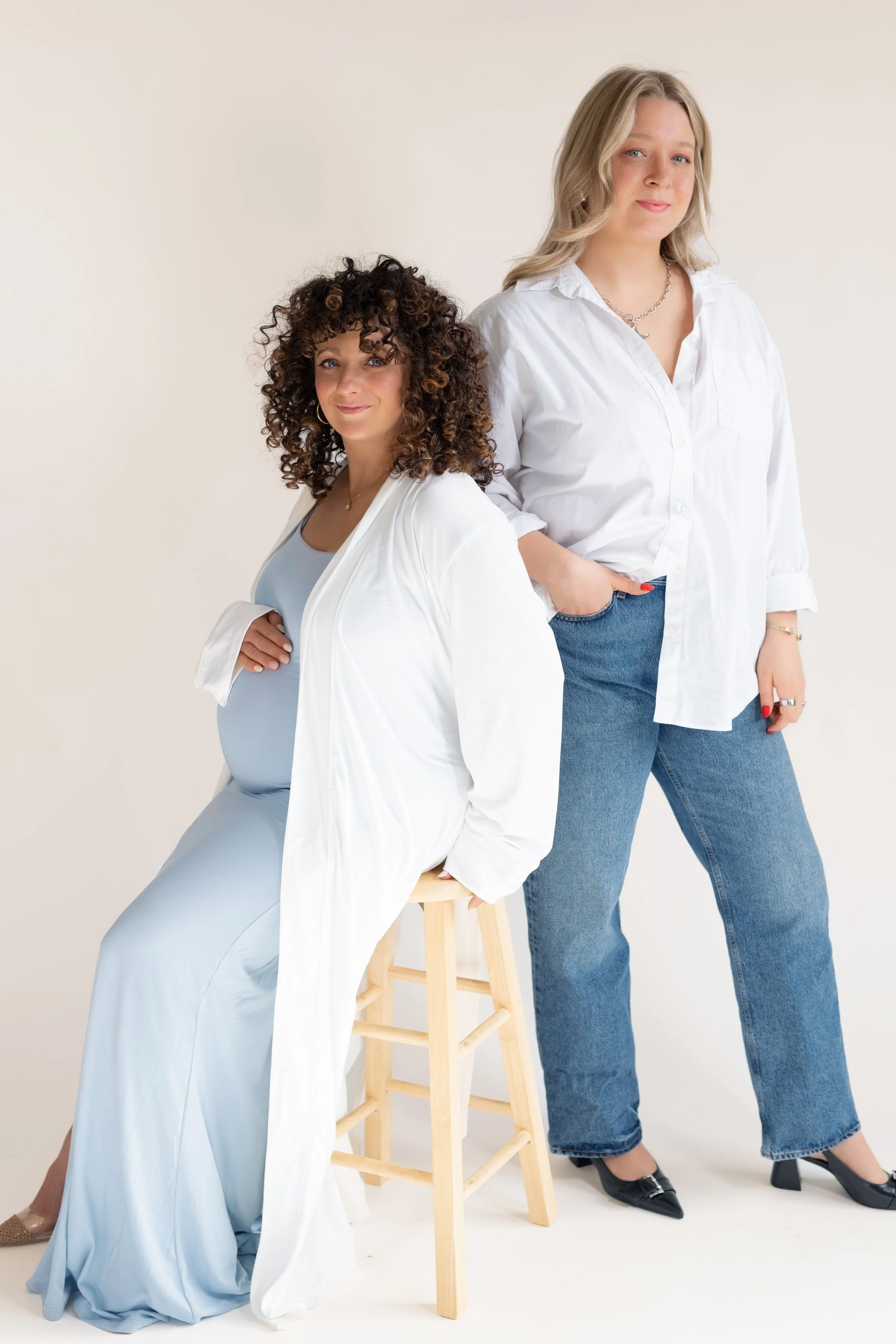 Two women posing in white tops and jeans against a light background. One is sitting on a stool with a light blue dress and the other is standing with her hand in her pocket.