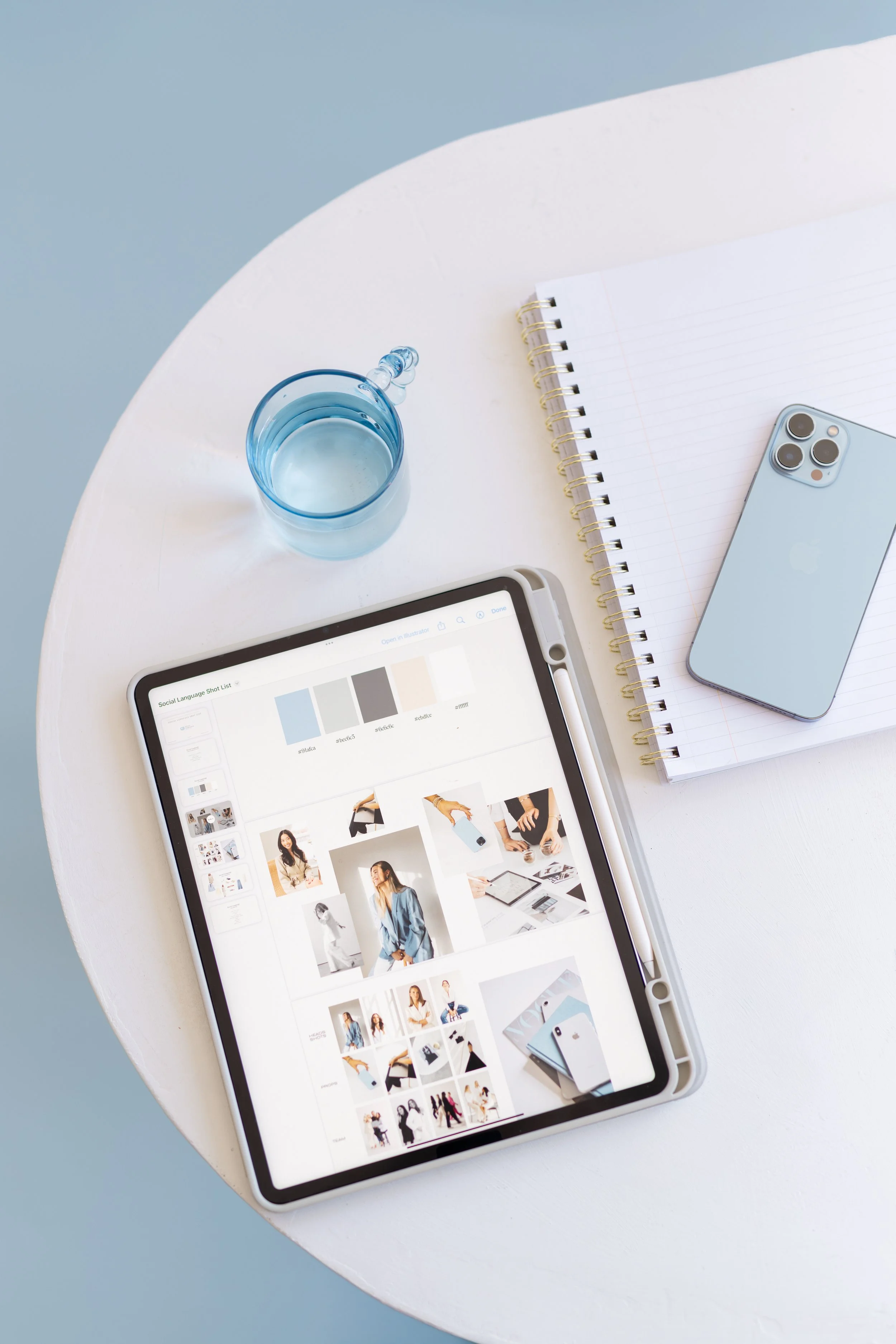 A tablet displaying a digital mood board of fashion and lifestyle photos, placed on a white table with a blue glass of water, a spiral notebook with lined paper, and a light blue iPhone.