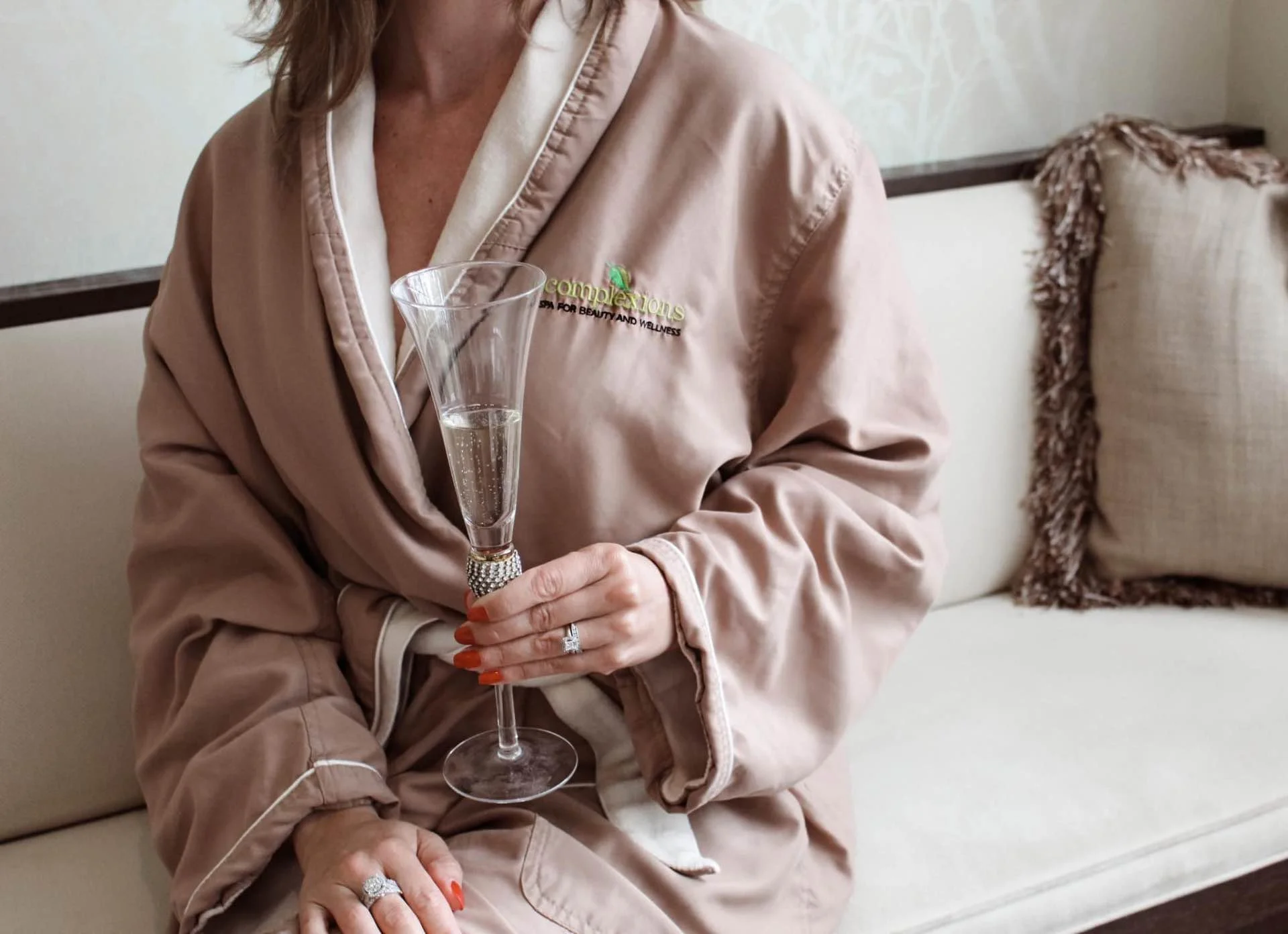 A woman wearing a beige robe with the logo 'complexions' embroidered on it, sitting on a cream-colored sofa holding a glass of champagne.