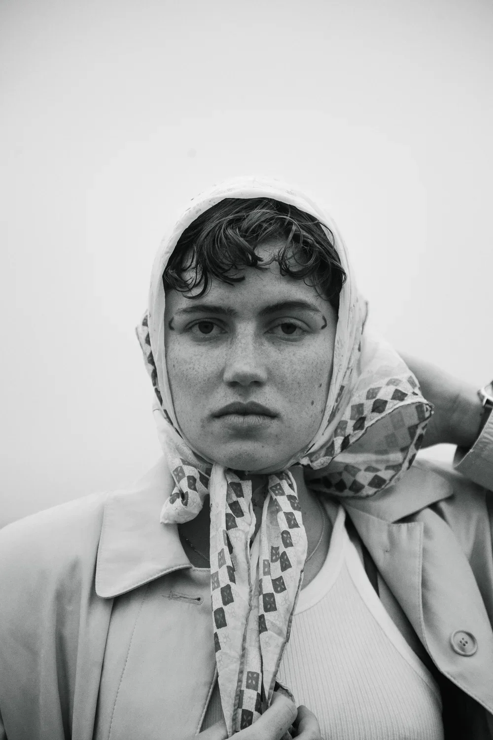 A black-and-white portrait of a person with short, wet hair, wearing a patterned headscarf, a light-colored trench coat, and a white top, looking directly at the camera against a plain background.