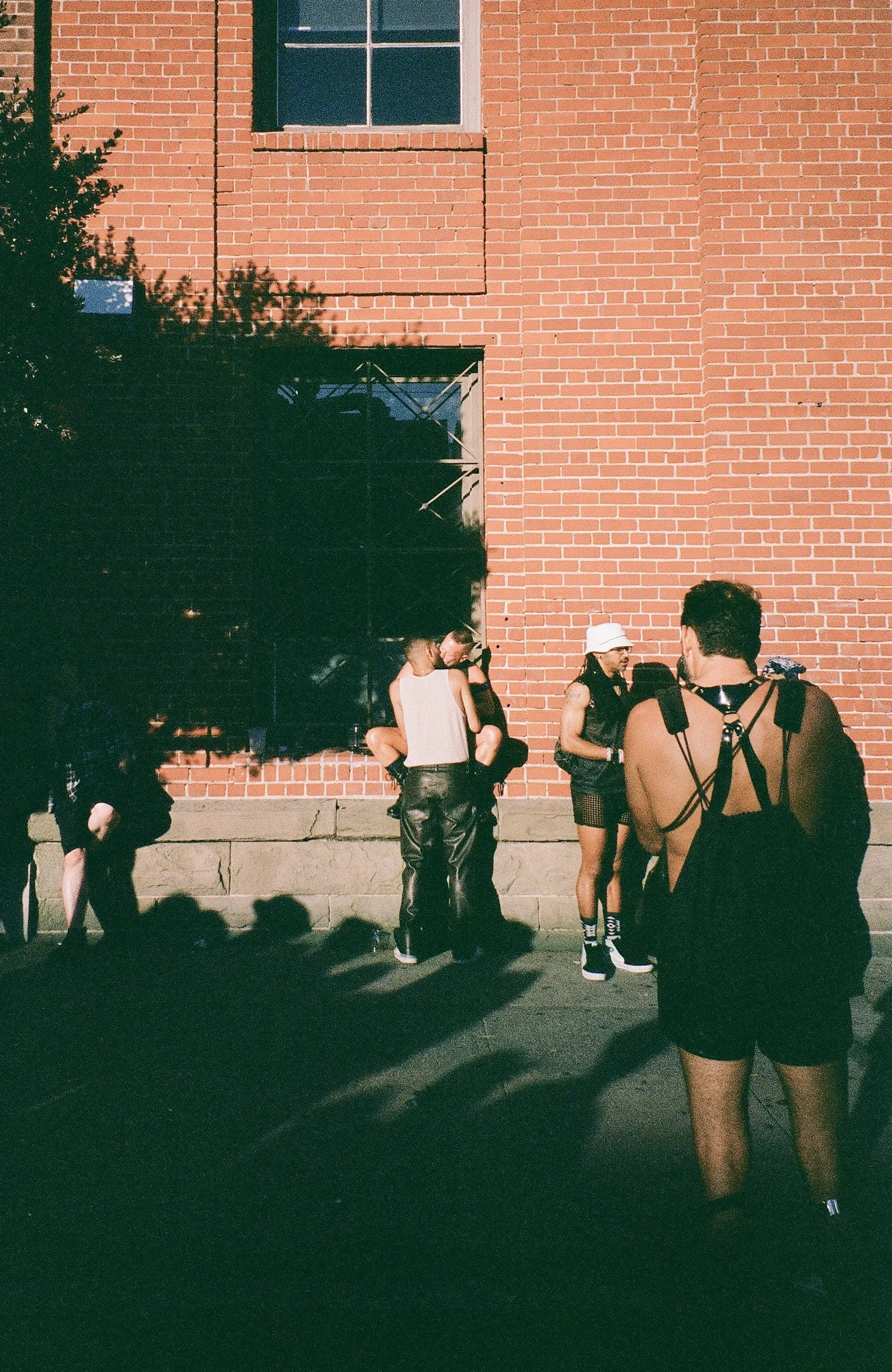 Group of people standing and sitting against a red brick wall, some dressed in alternative fashion, in sunlight with shadows on the ground.