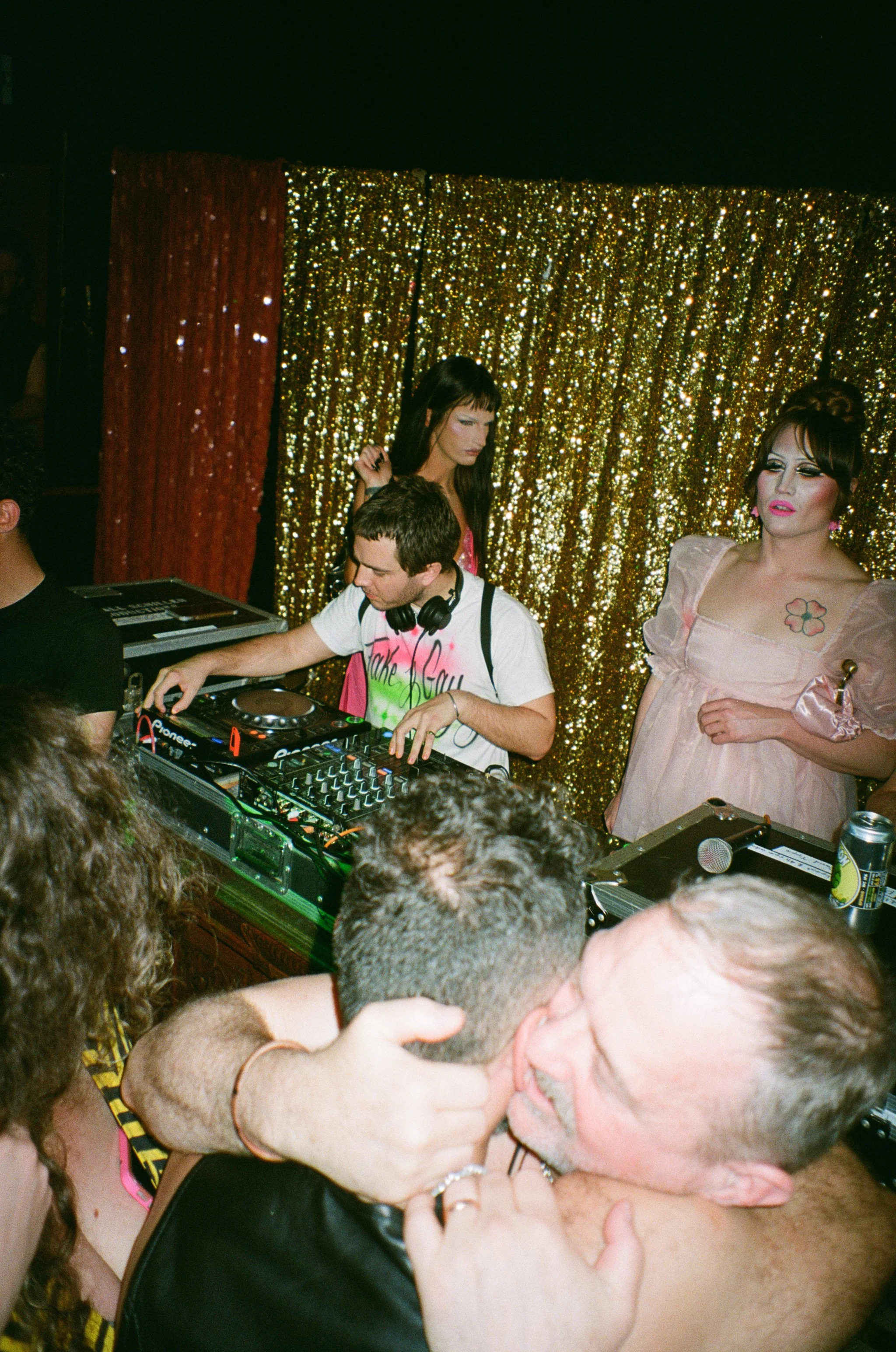 A DJ is mixing music at a party with a gold sequin backdrop. Two women in pink dresses stand behind him; one has dark hair and the other has makeup and a tattoo. In the foreground, two men are dancing closely, celebrating.