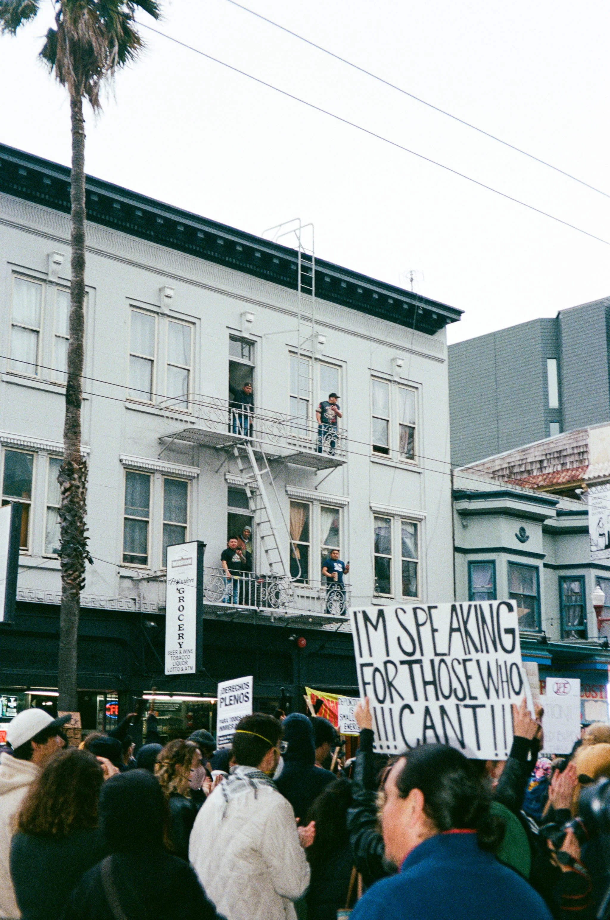 People protesting on a street in front of a white building with several individuals on the balconies holding signs. The protester is holding a sign that reads, 'I'M SPEAKING FOR THOSE WHO CAN'T!!!'